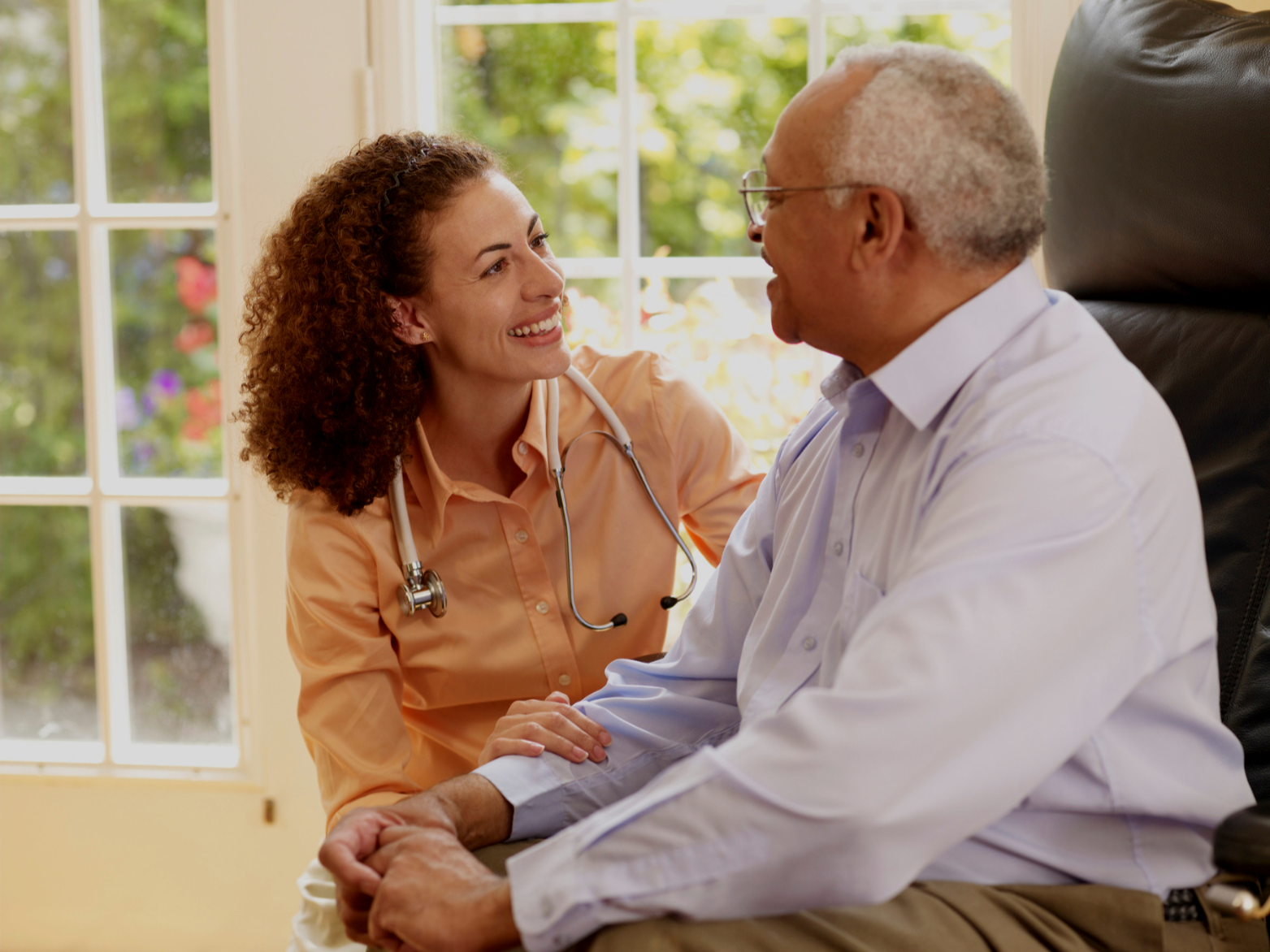 A young woman healthcare professional in an orange shirt with a stethoscope around her neck, smiling and holding the hand of an elderly man in a white shirt, sitting in a chair near a window with a garden outside.