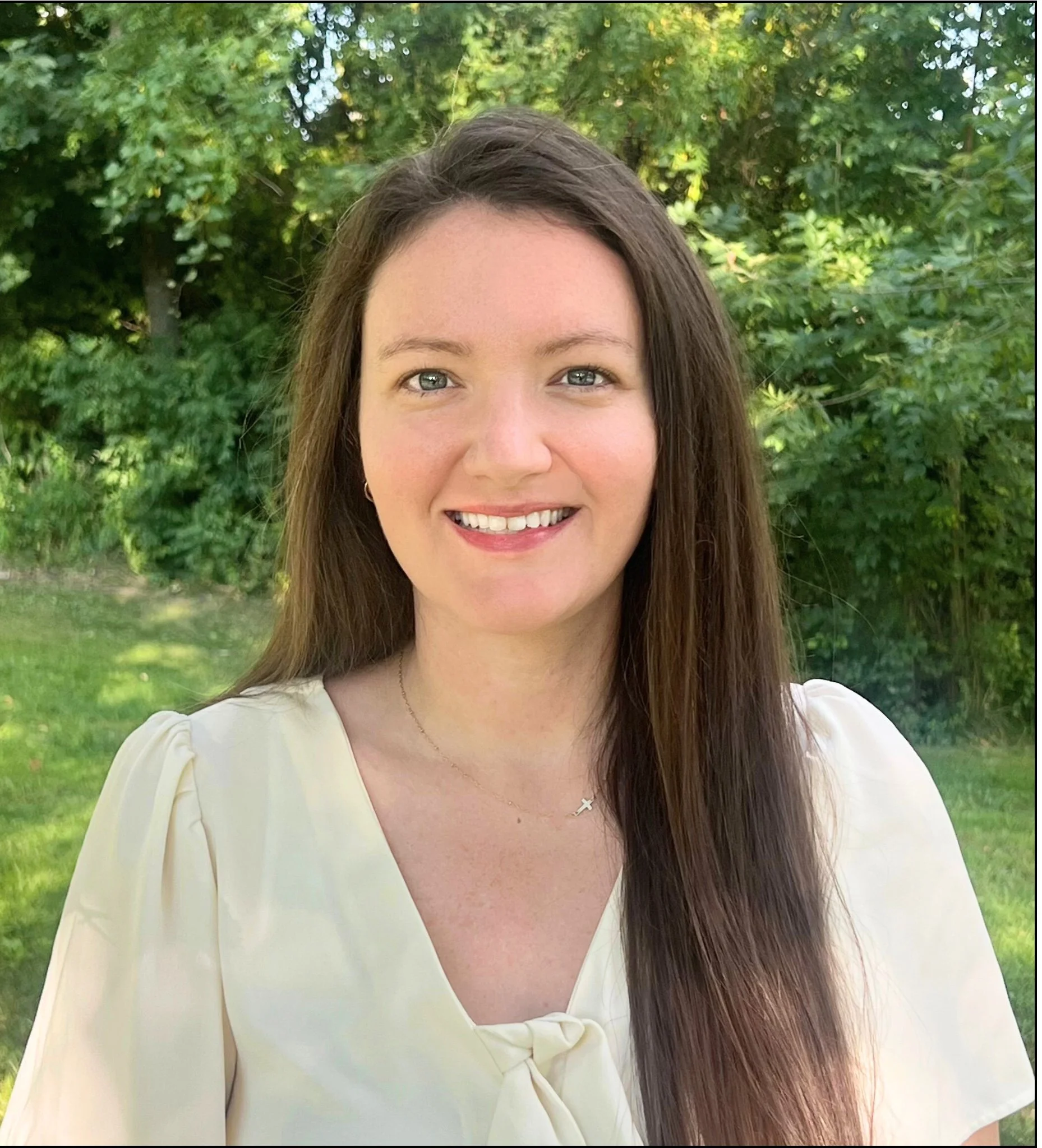 A woman with long brown hair smiling outdoors in front of green trees, wearing a light yellow blouse and a delicate necklace.