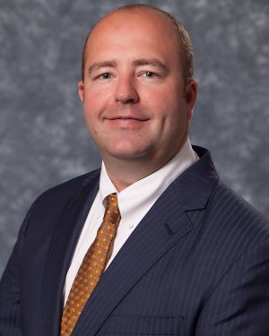 Professional portrait of a man in a suit with a white shirt and tie, smiling against a gray background.