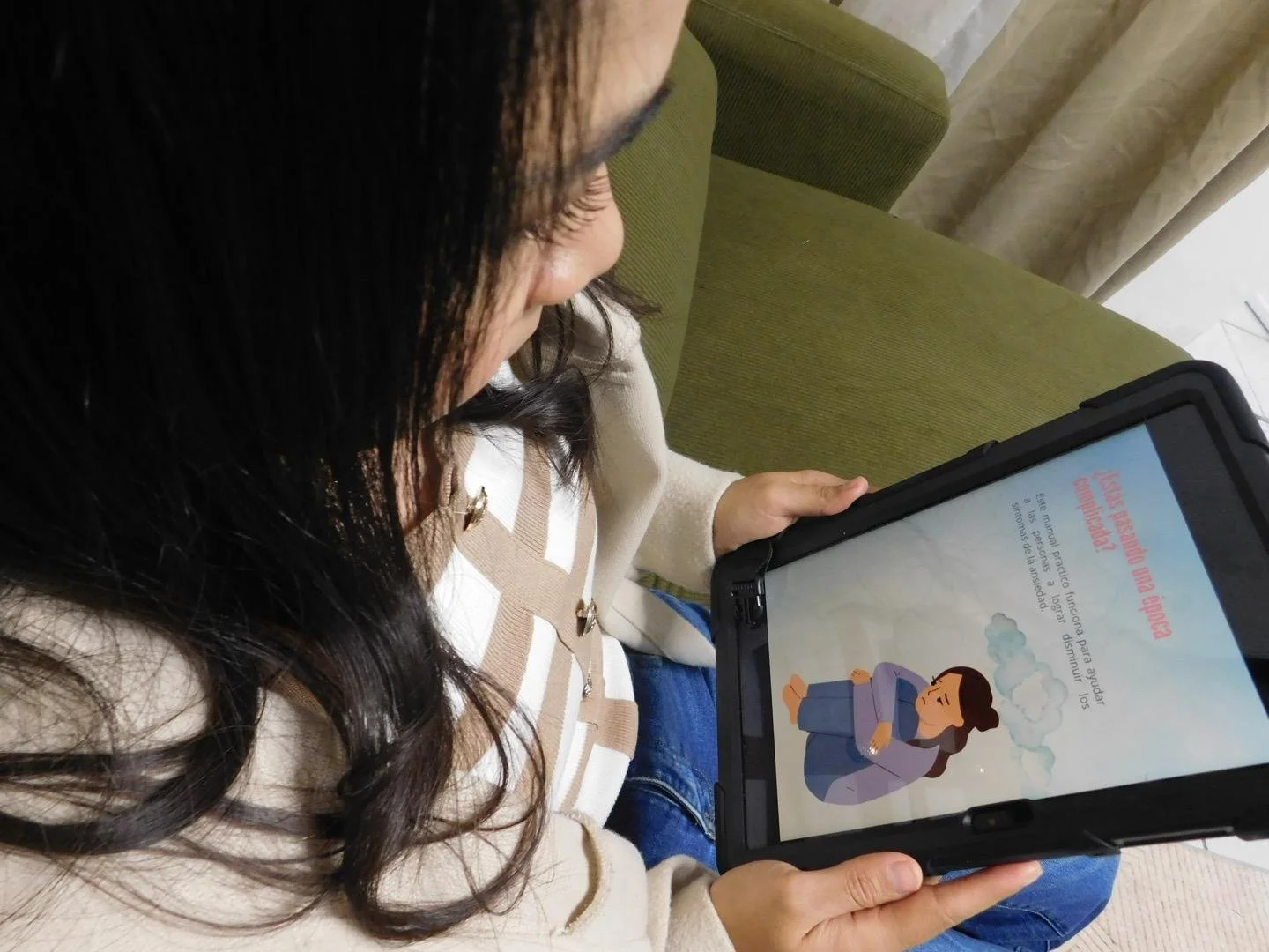 Una mujer con cabello largo oscuro lee en una tableta que muestra un dibujo y texto en español en una sala con sofá verde y cortinas beige.