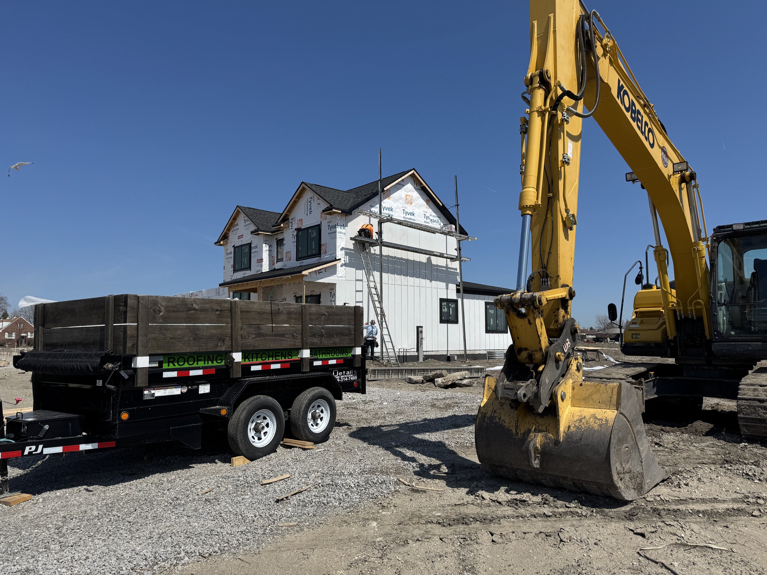 Construction site with a house under construction, a yellow excavator, and a trailer labeled for roofing, kitchens, and bathrooms, on a gravel lot under a clear blue sky.