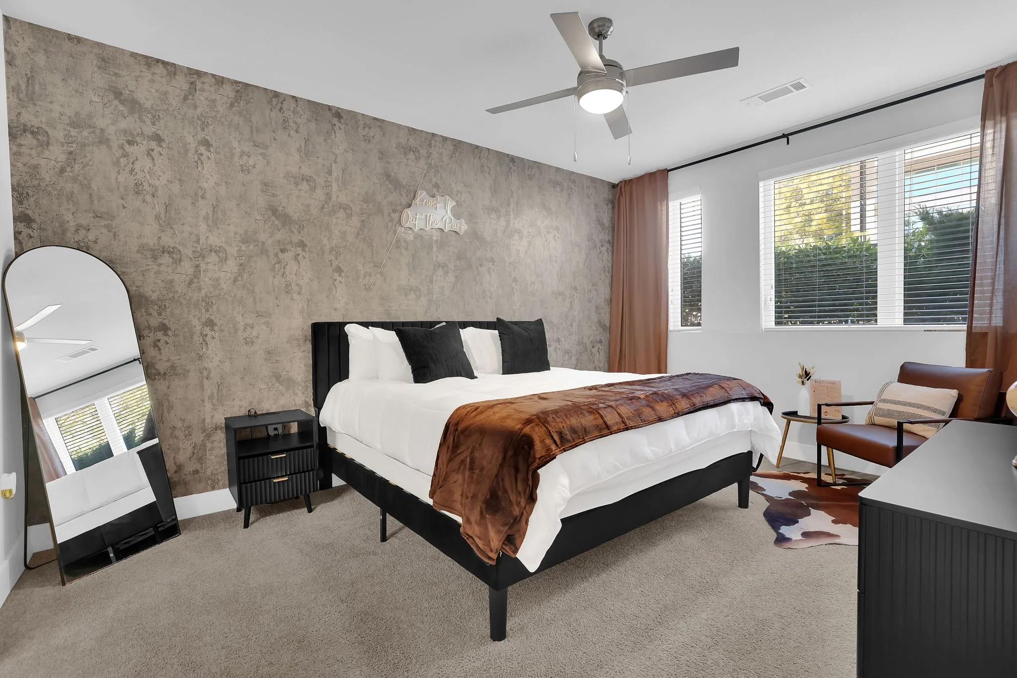Modern bedroom with a large bed, black headboard, white bedding, brown throw blanket, and black pillows. Includes a black nightstand, a full-length mirror, a brown chair, and a window with blinds and curtains. Pendant light and ceiling fan.