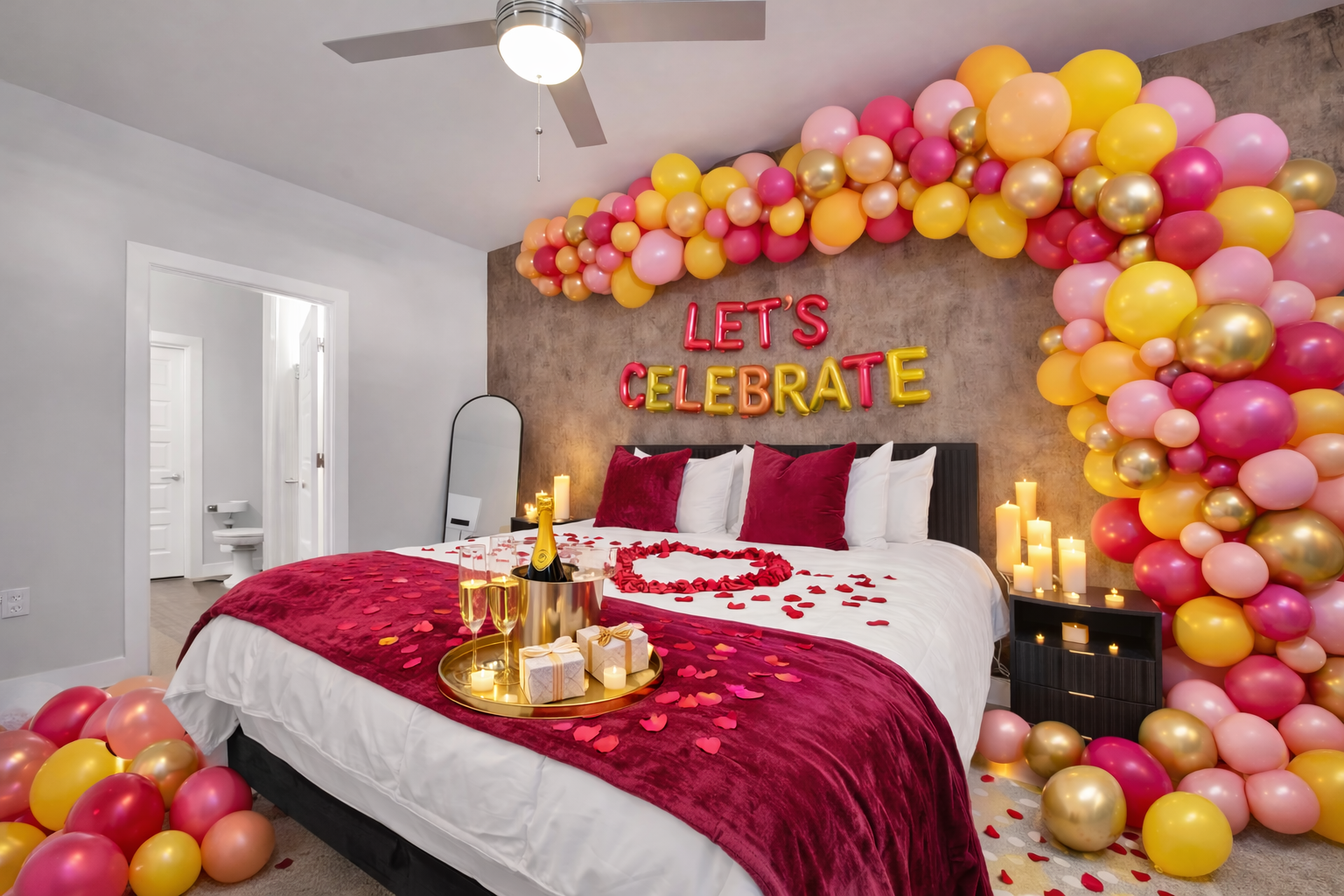 Decorated bedroom with loop balloon arch, candles, and 'Let's Celebrate' sign over the bed, rose petals, and celebratory items on the tray.