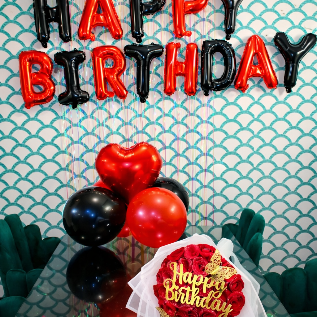A birthday celebration setup featuring red and black balloon letters spelling out "HAPPY BIRTHDAY," with additional red and black balloons, a bouquet of red roses with a gold "Happy Birthday" sign and butterfly decorations, and a decorative blue and 