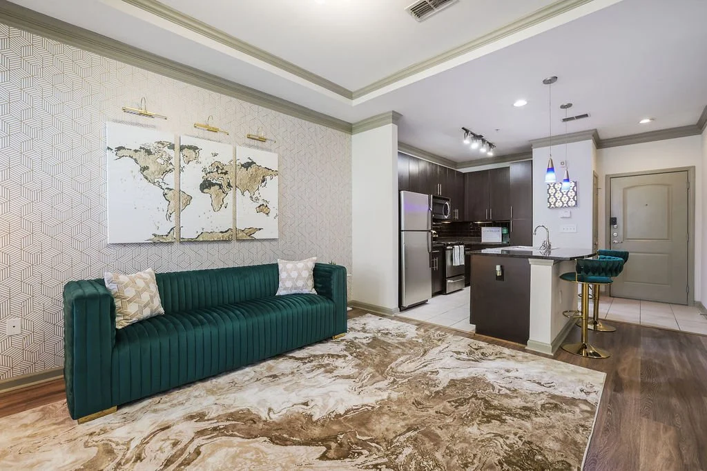 Living room with green velvet sofa, world map artwork, beige patterned wallpaper, marble-patterned rug, open kitchen with dark cabinets, stainless steel appliances, and small bar stools.