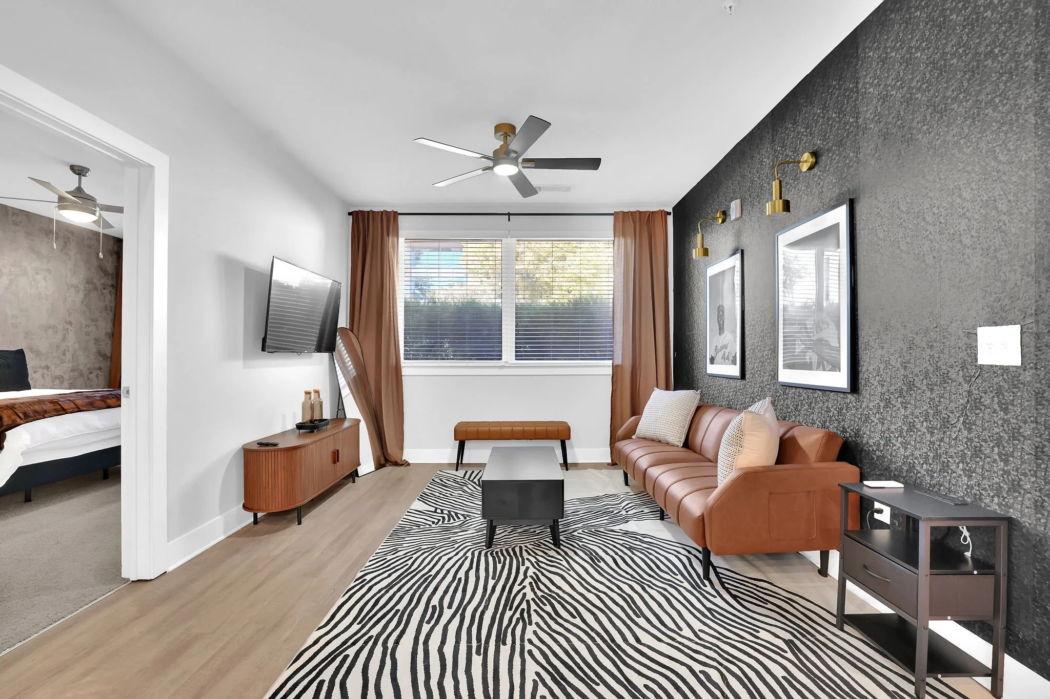 Living room with a black textured accent wall, a tan leather sofa, black and white zebra print rug, black coffee table, wall-mounted TV, window with brown curtains, and framed black-and-white artwork with gold lighting fixtures.
