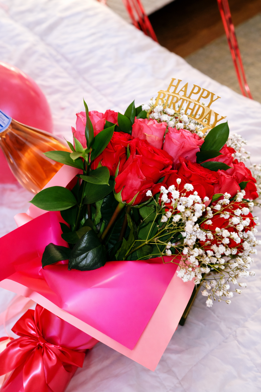 Bouquet of pink and red roses, white baby's breath, and green leaves with a gold 'Happy Birthday' sign, wrapped in pink paper with a large red ribbon, on a white tablecloth.