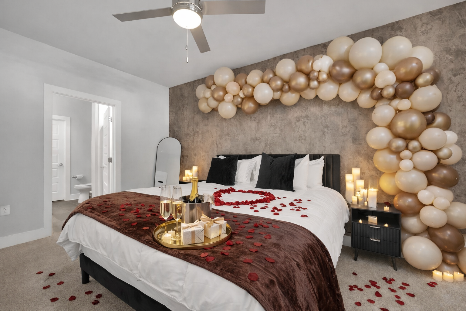 A romantic bedroom decorated with balloons, candles, and rose petals. The bed has a dark headboard, white bedding, red rose petal hearts, and two black pillows. A gold tray with champagne, glasses, and gifts is on the bed. The wall behind the bed has