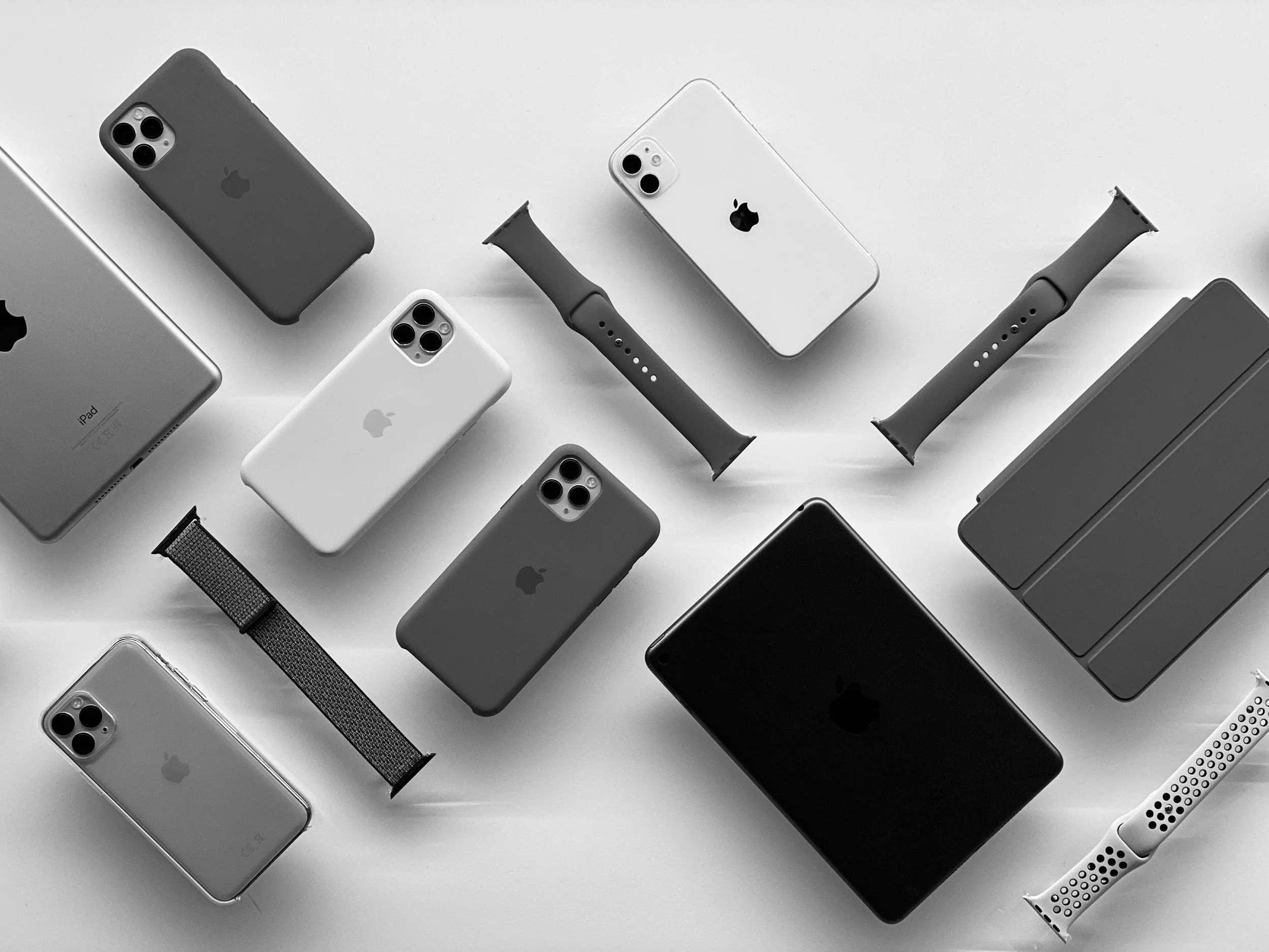 Multiple Apple devices including iPhones, iPads, Apple Watches, and accessories arranged on a white surface in black and white.