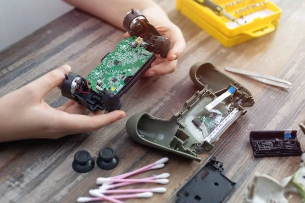 Person repairing a gaming controller with its top shell removed, exposing the internal circuit board. Tools and small parts are laid out on a wooden table.