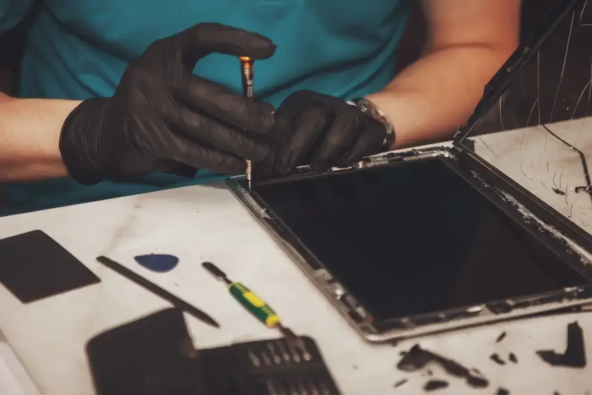 Person wearing black gloves repairing or assembling a device, possibly a tablet or phone, with a screwdriver, on a cluttered workbench.