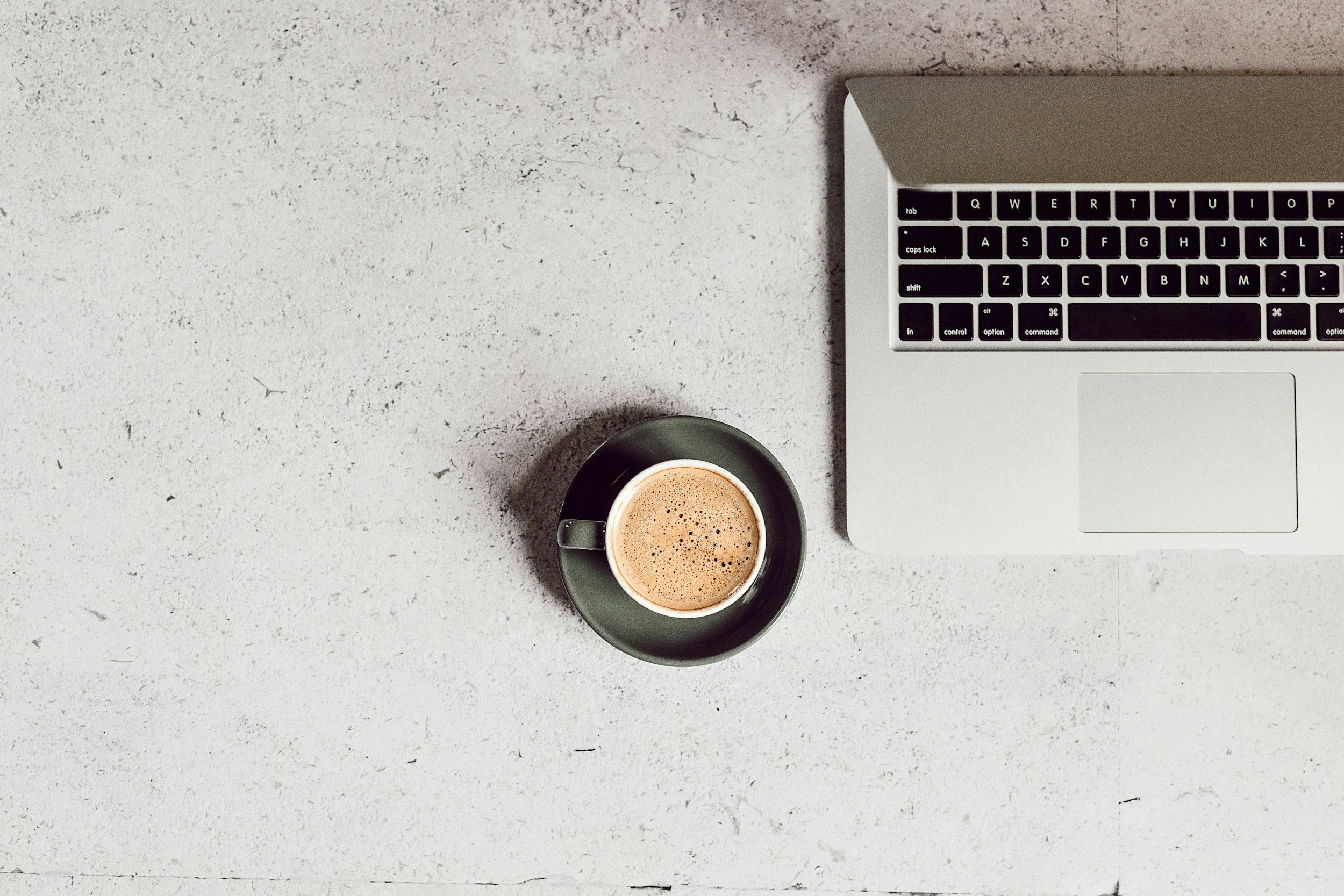 Top view of a workspace with a silver laptop and a black coffee cup on a white textured surface.