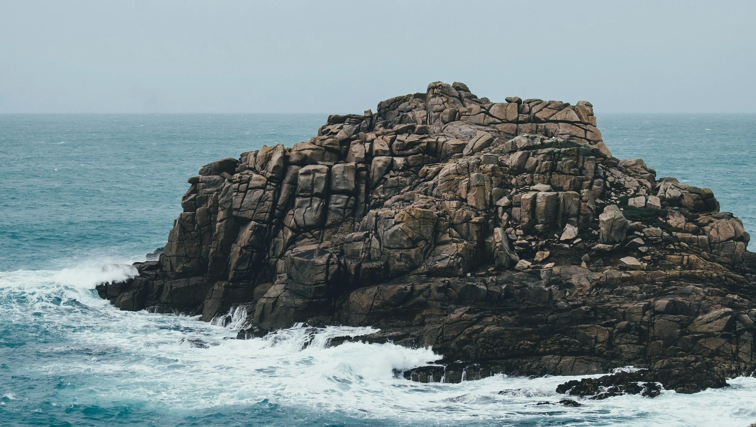 A large rocky formation in the ocean with waves crashing against it, under a cloudy sky.
