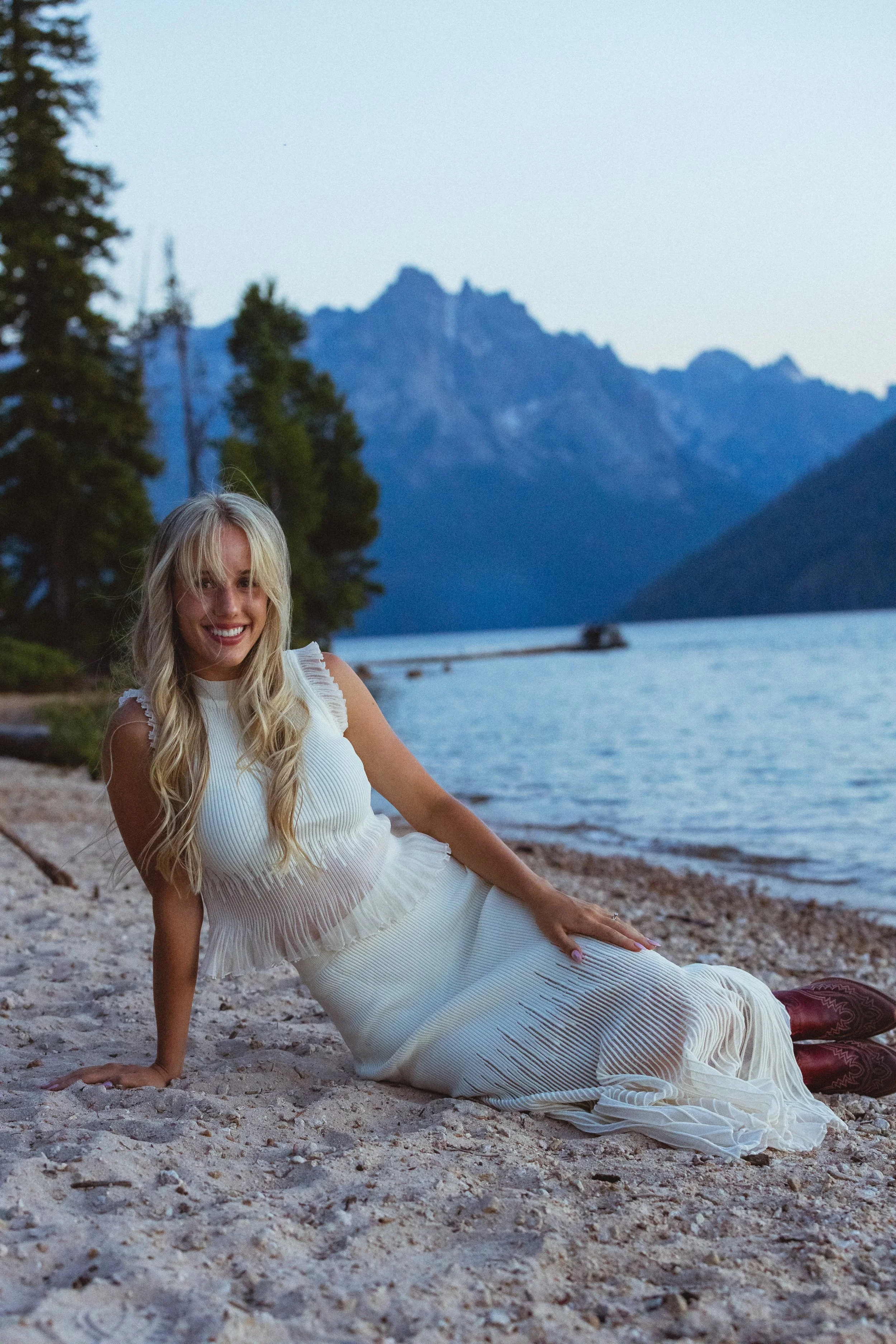 A woman in a white dress is sitting on a sandy beach near a lake with mountains and trees in the background, smiling at the camera.