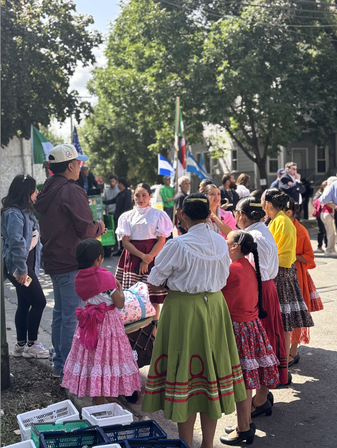 People dressed in traditional Mexican attire gathered outdoors at a cultural event with flags in the background.