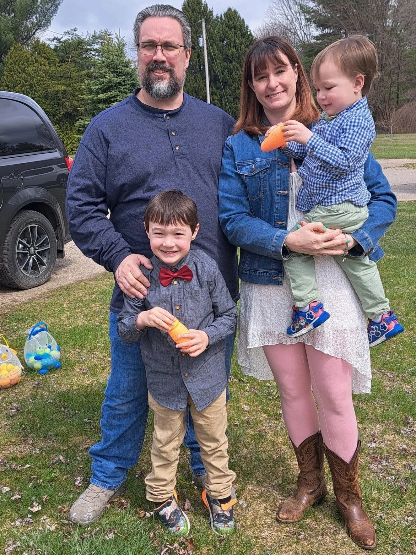 A family of four standing outdoors in a grassy area with trees in the background. The father has gray hair and a beard, wearing glasses and a navy blue shirt. The mother has red hair, wearing a denim jacket. The son, standing in front of his father, is smiling, wearing a gray shirt with a red bowtie and khaki pants. The younger child, being held by the mother, is holding a carrot and wearing a blue checkered shirt, beige pants, and colorful shoes. There are Easter egg baskets on the ground nearby.