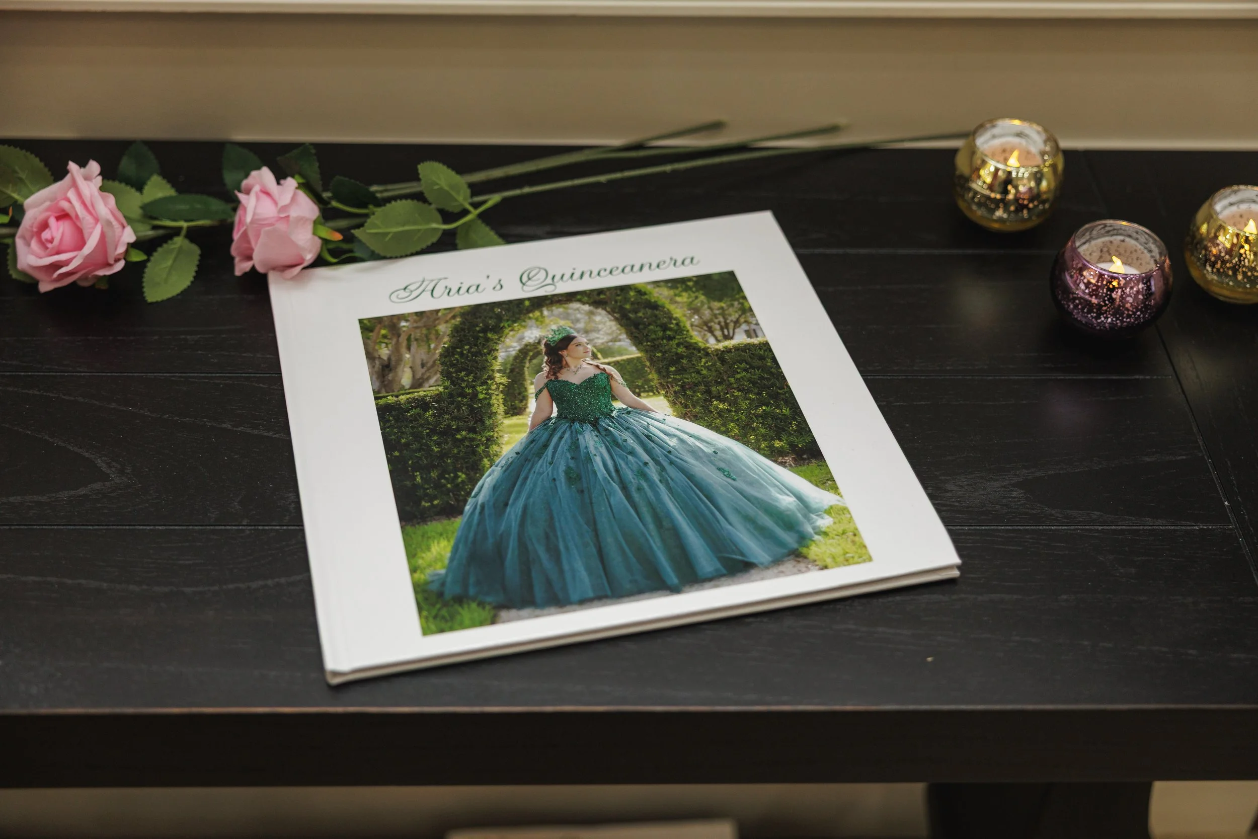 A photo album titled 'Aria's Quinceanera' on a wooden table, showing a girl in a green and blue quinceañera dress, surrounded by pink roses and lit candles.