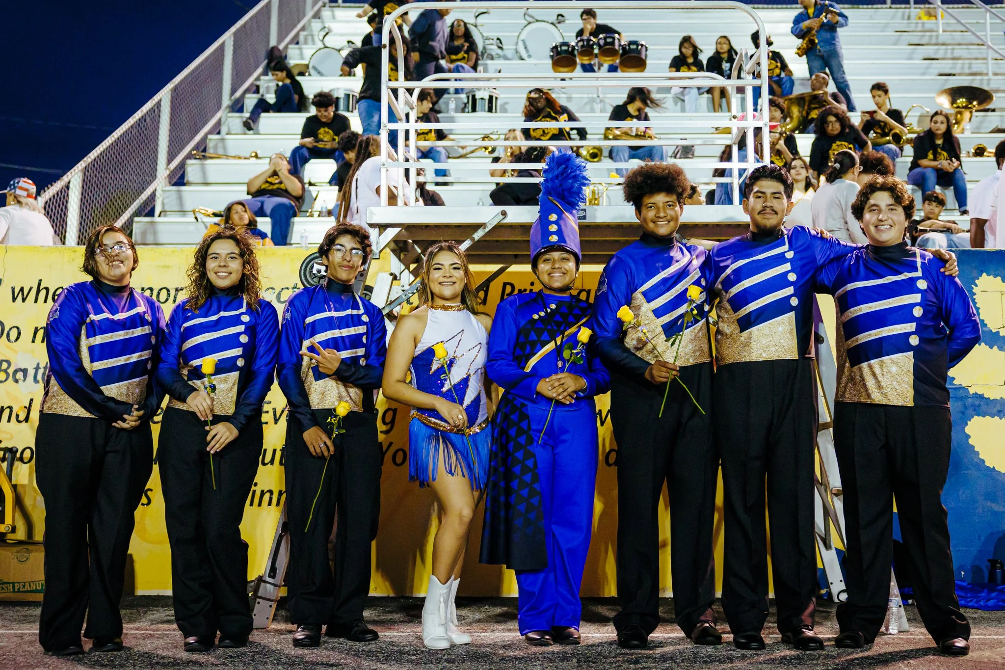Group of people in marching band uniforms standing together on a football field, holding yellow roses, with band members and drums in bleachers behind them.