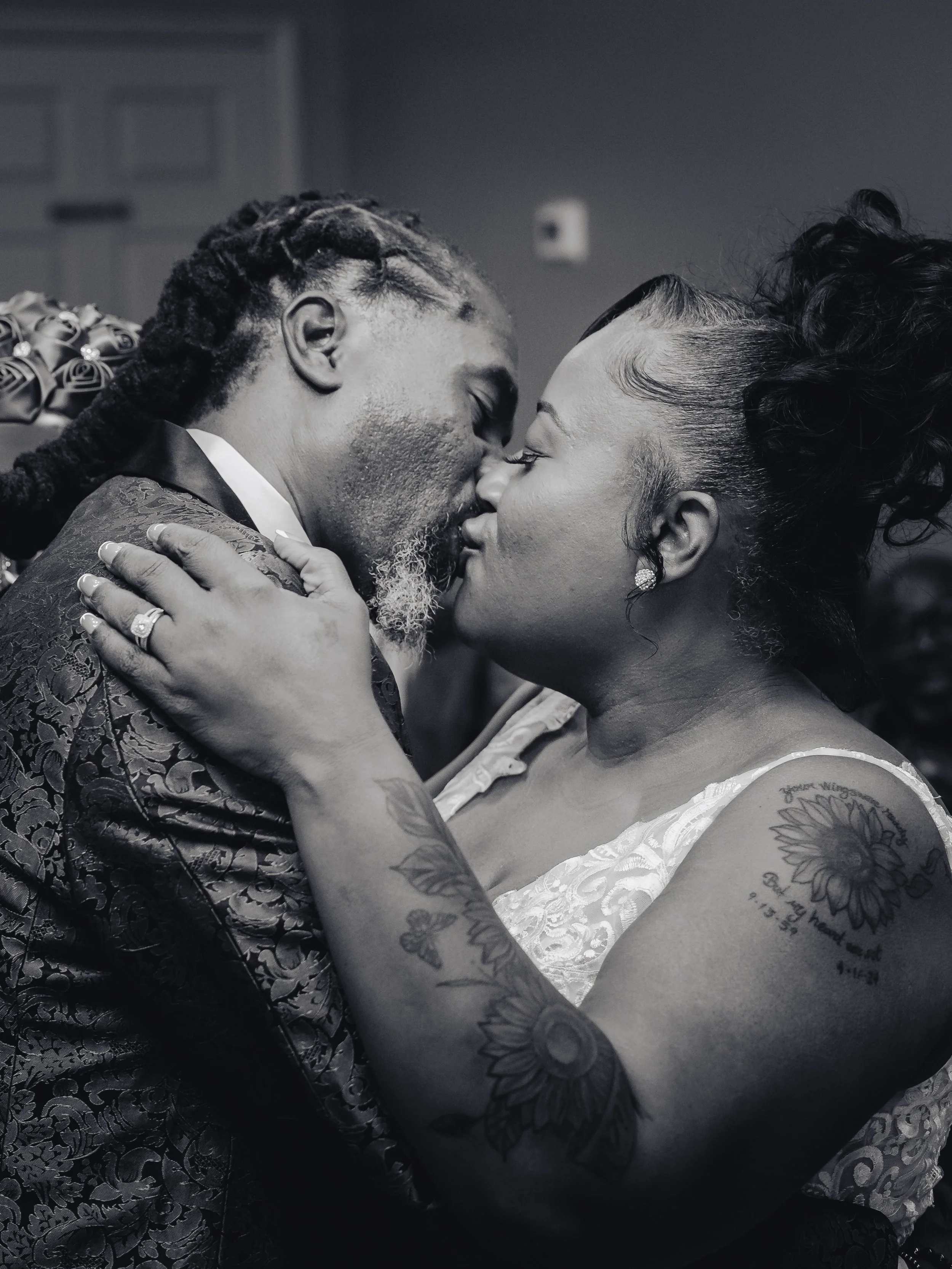 A black-and-white photo of a couple embracing and kissing at a wedding. The man has braided hair and a beard, wearing a floral-patterned jacket, and the woman has her hair styled up, with tattoos on her arm and shoulder, wearing a lace dress.