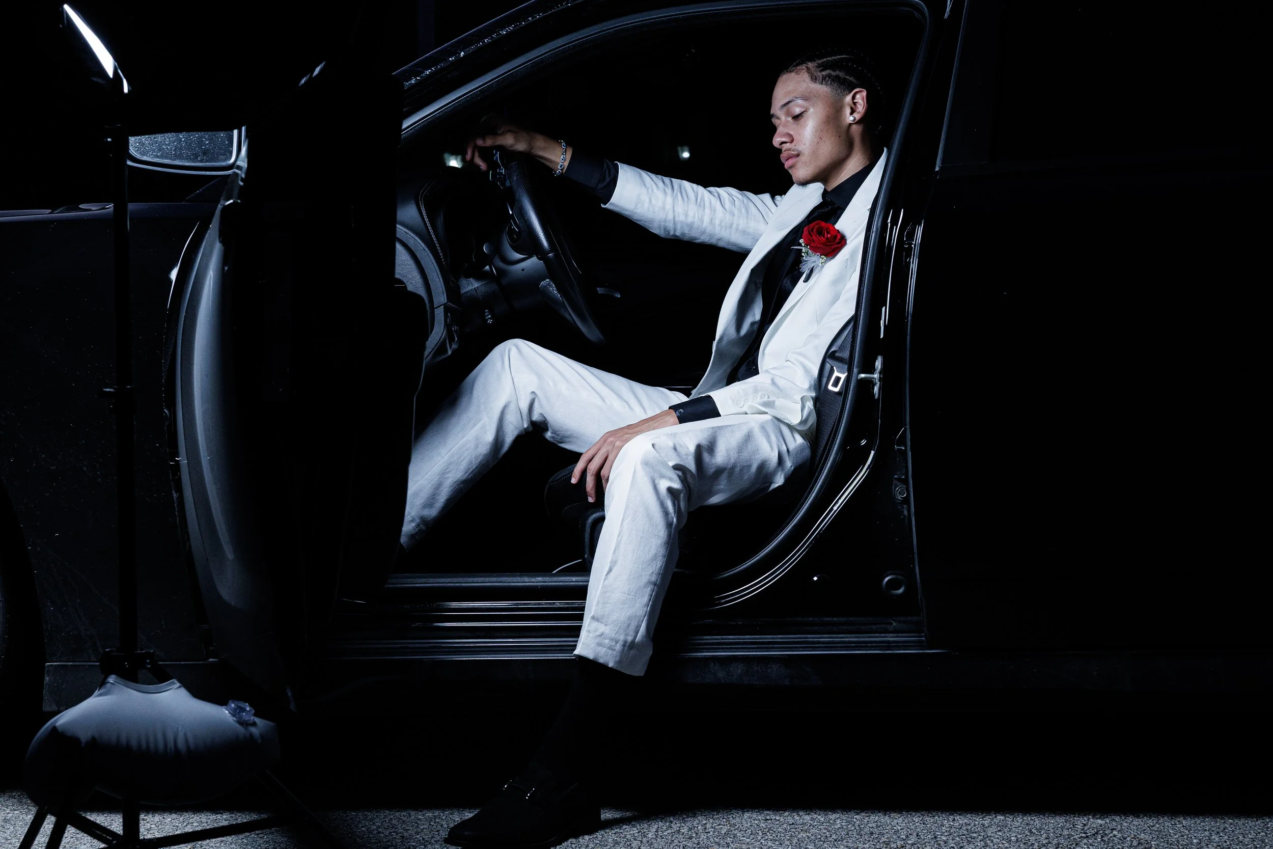 Young man sitting in the driver seat of a car at night, dressed in a white suit with a red rose on his lapel, looking down with his eyes closed.
