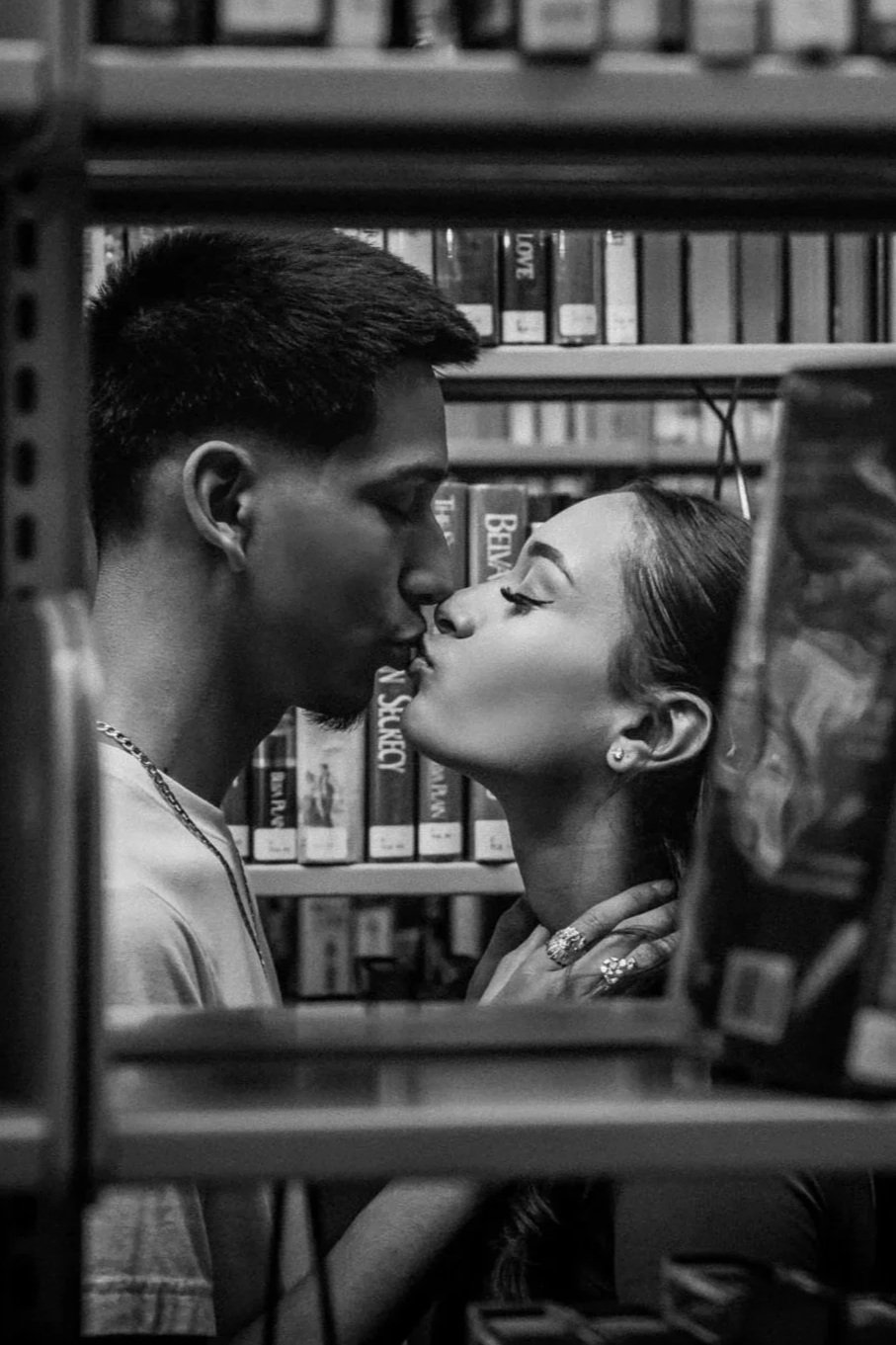A black and white photo of a young couple kissing in a library, surrounded by shelves of books.