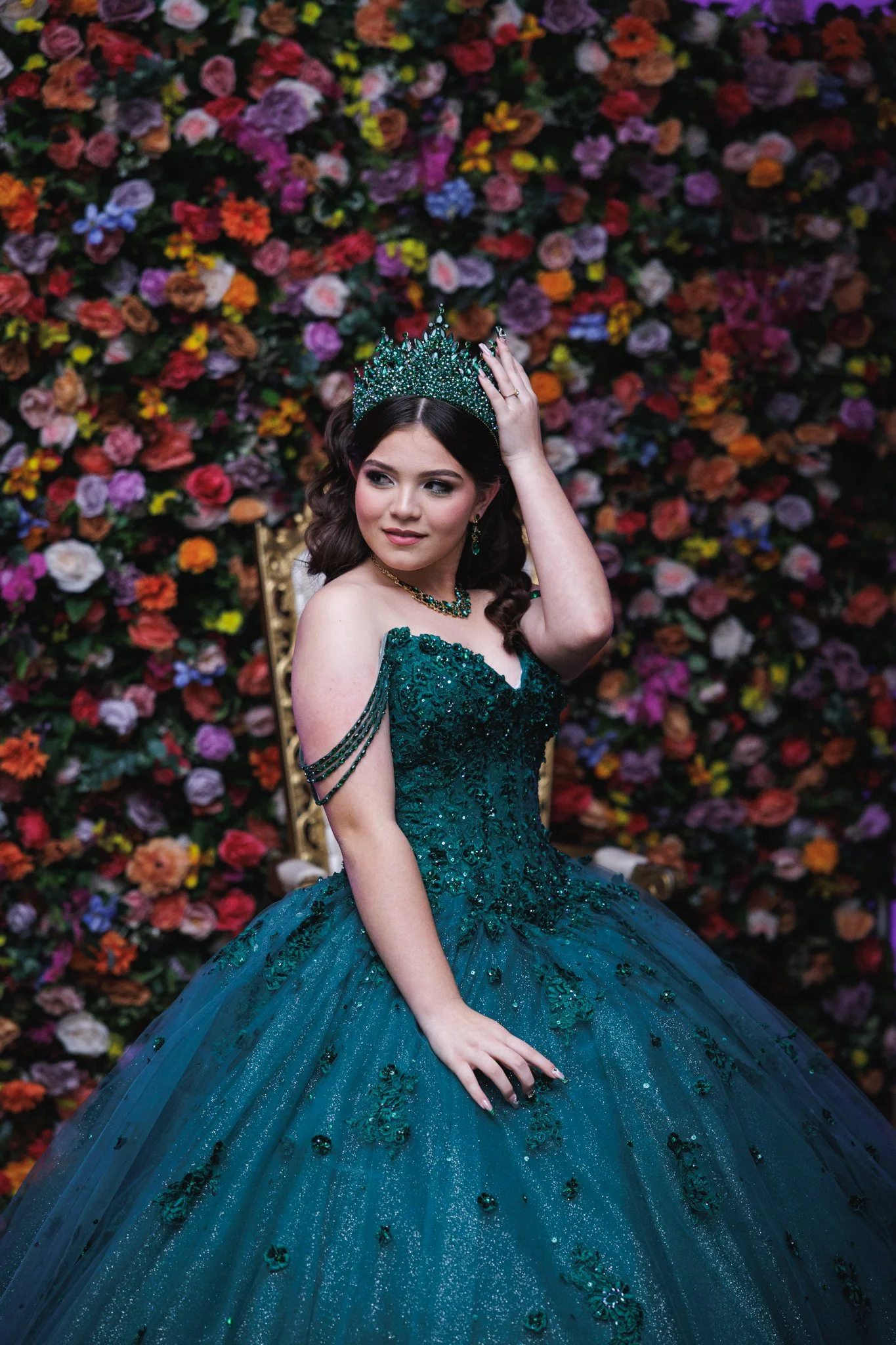 A young woman in an elaborate green ball gown with beaded details and off-shoulder straps, wearing a matching crown and jewelry, sits on a golden throne in front of a backdrop filled with colorful flowers.