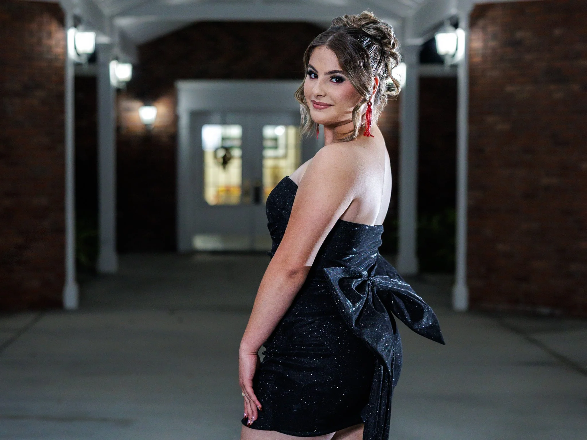 A young woman in a black strapless dress with a large bow at the back, standing outdoors at night near a building with brick walls and white trim, smiling and looking over her shoulder.