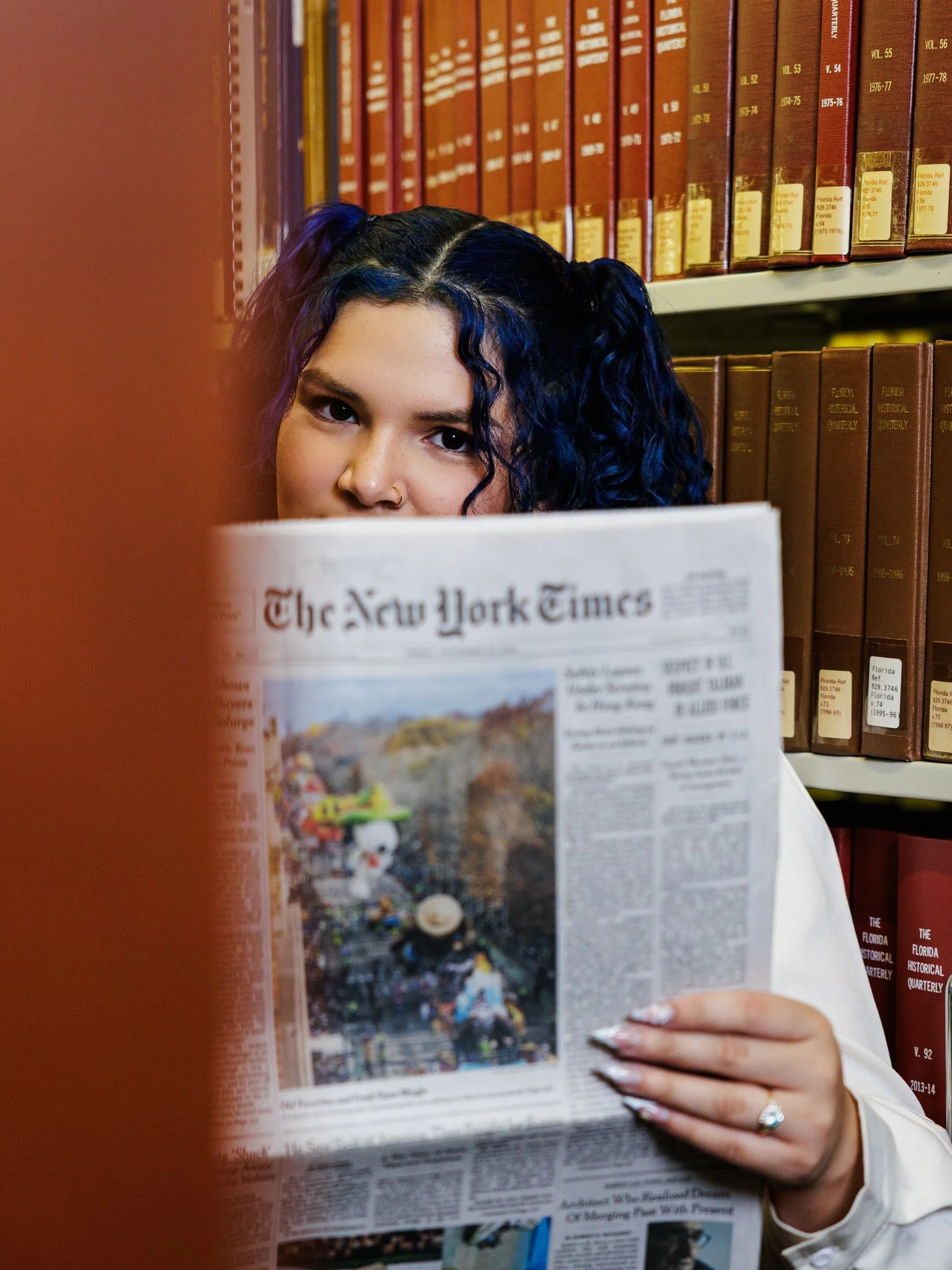 A young woman with blue curly hair and a nose ring reading a newspaper in a library, with shelves of brown and red books in the background.