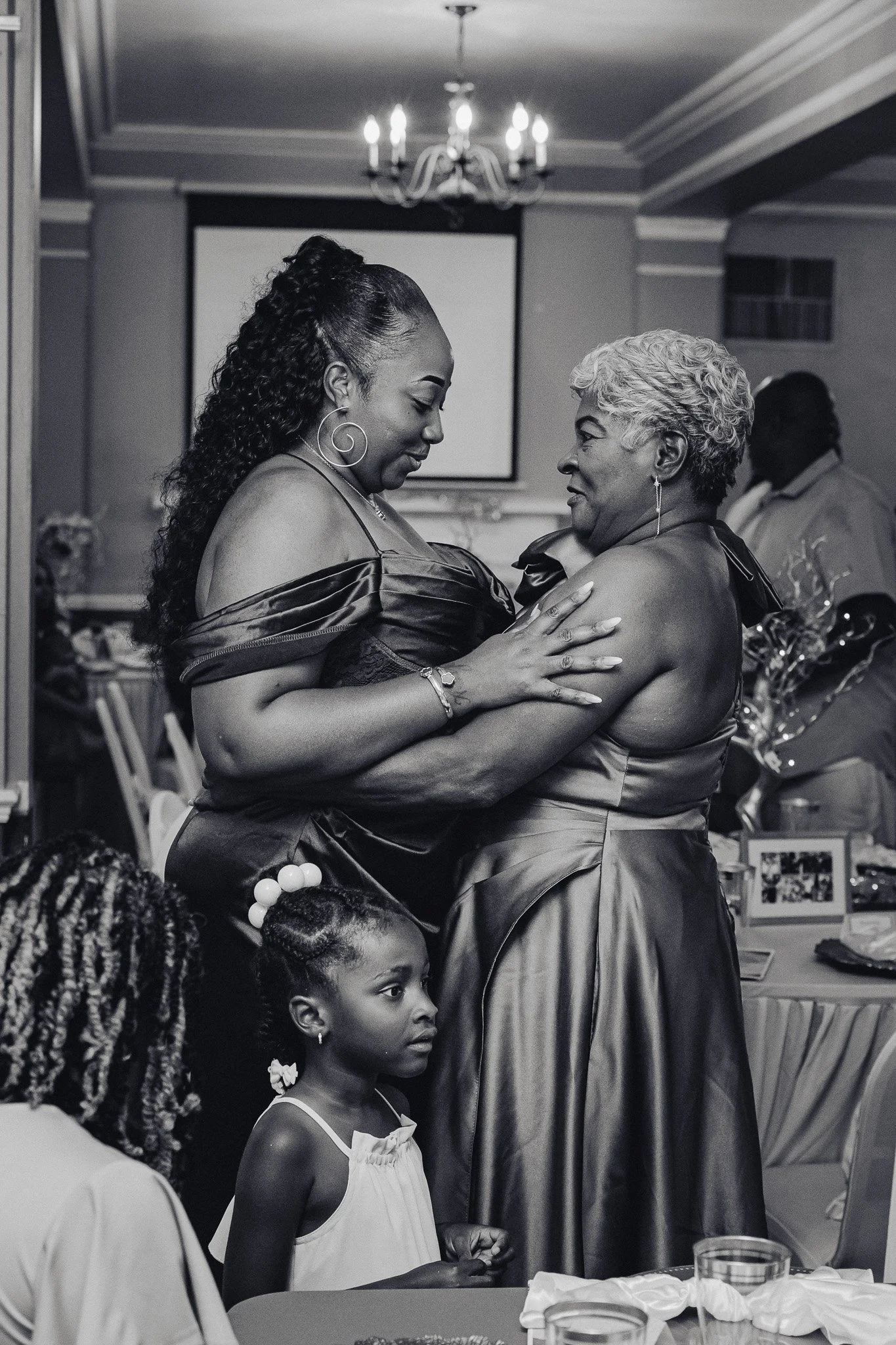 Two women, one older and one younger, are dancing close together in a celebration setting, with children and other guests watching nearby.