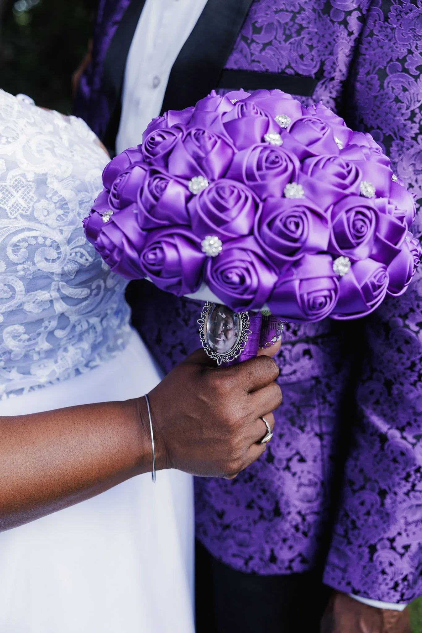 A close-up of a wedding bouquet made of purple satin roses with decorative rhinestones, being held by a woman wearing a lace wedding dress, with a man in a purple patterned jacket in the background.