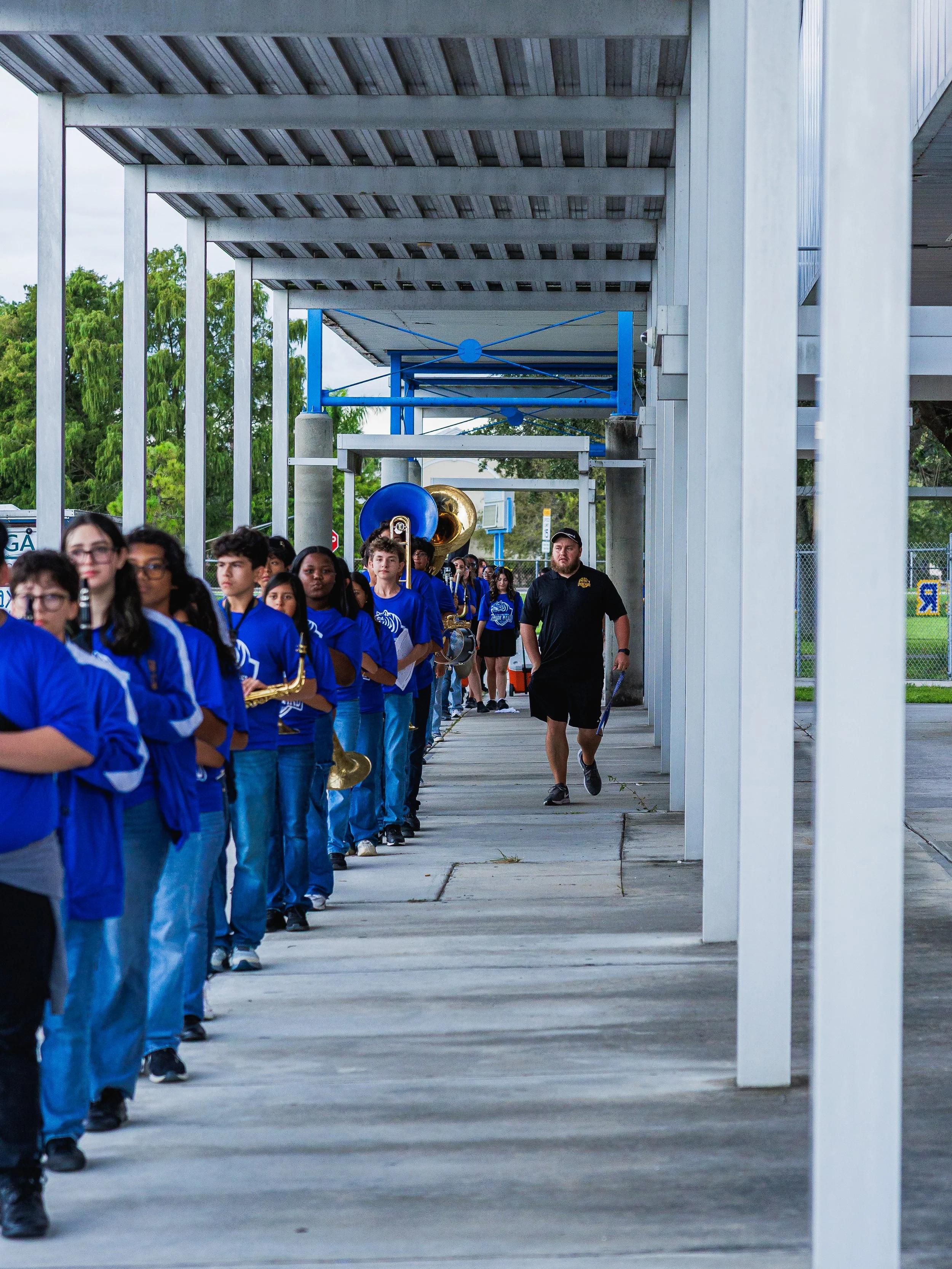 A line of students wearing blue shirts and jeans, holding musical instruments, walking under a covered walkway at a school, led by a man in black walking alongside.