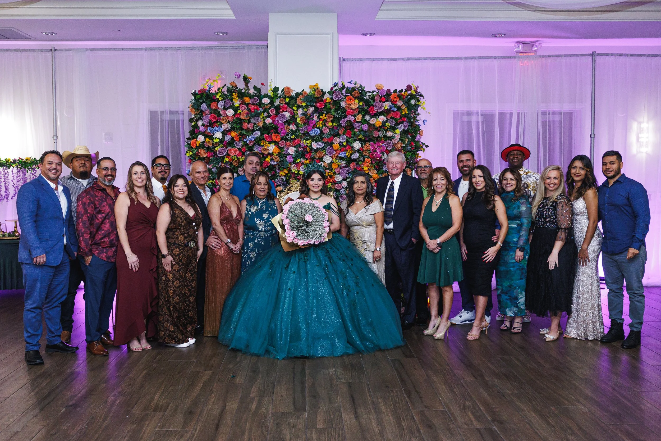 Group of people at a celebration, with a woman in a teal ball gown holding a bouquet of flowers, standing in front of a colorful floral backdrop.