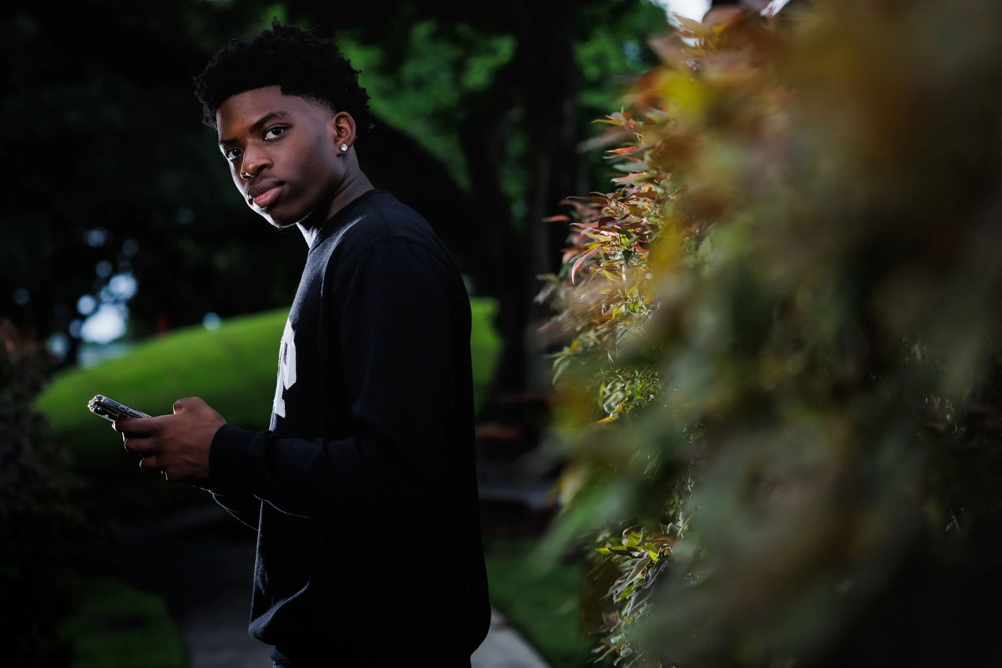 A young man with dark skin, curly black hair, wearing a black jacket, holding a smartphone, stands outdoors in a park with green trees and bushes.
