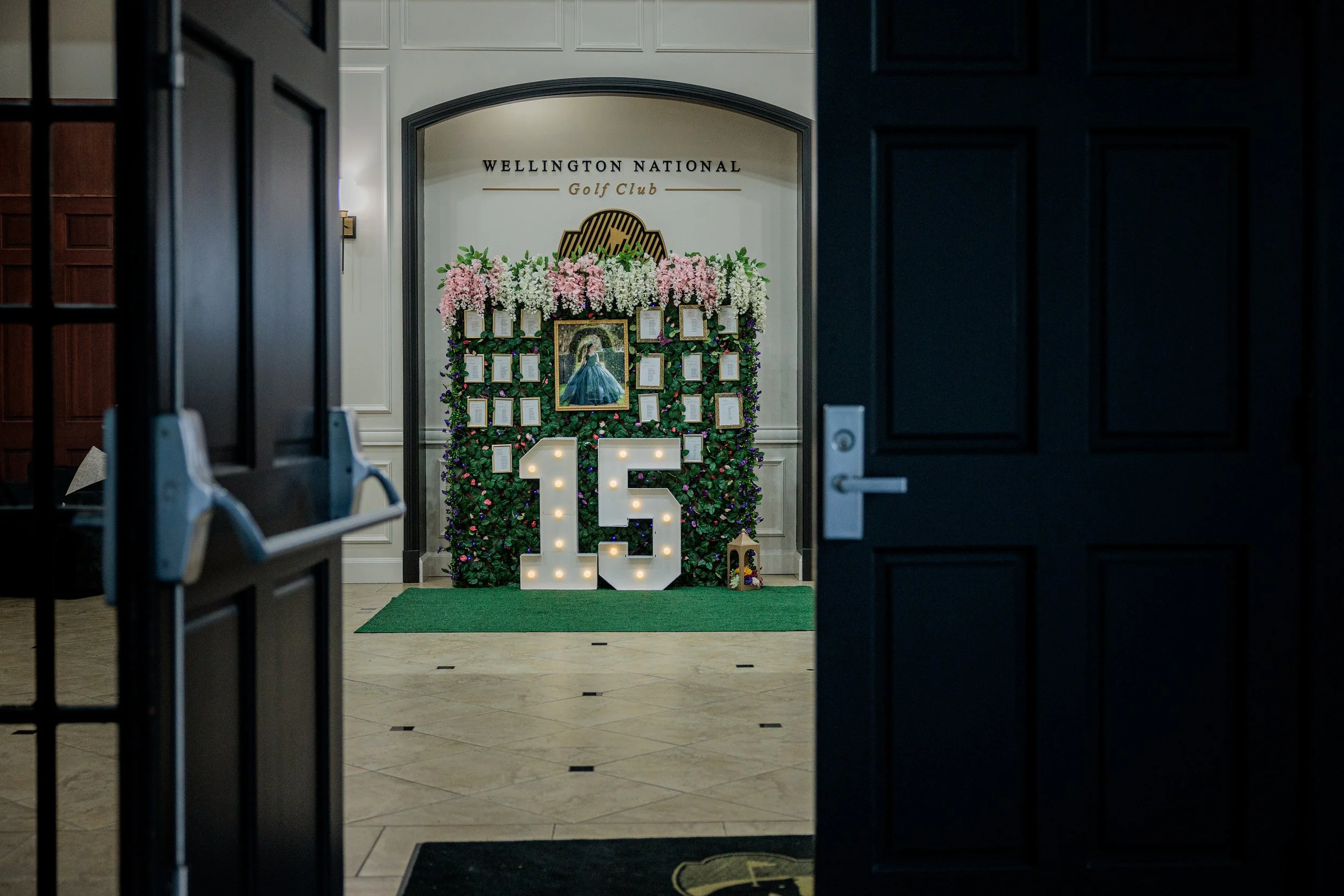 Decorative display at Wellington National Golf Club for a 15th anniversary celebration, featuring a floral backdrop, a framed portrait of a woman, an illuminated number 15, and a small nativity scene.