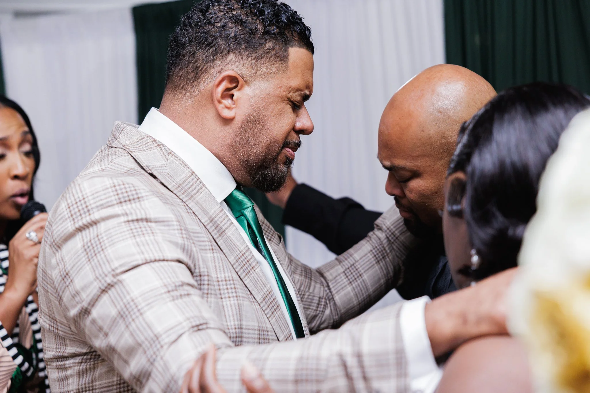A close-up of a man in a plaid suit with a green tie and a goatee praying with his eyes closed and hands placed on a person's shoulders. The person being prayed for appears to be kneeling with their head bowed. There are other individuals in the back
