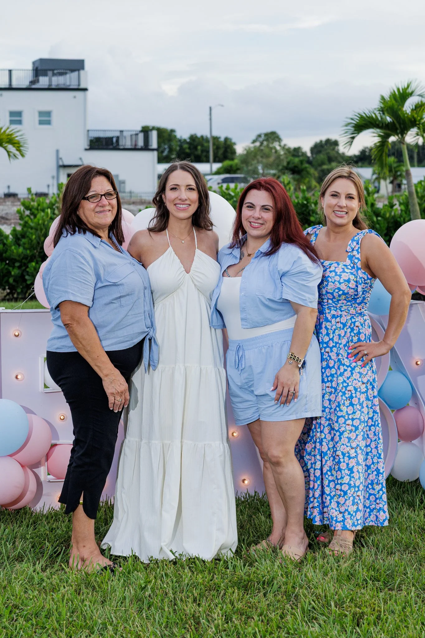 Four women standing outdoors in front of a pink and blue balloon display during daytime, smiling at the camera.