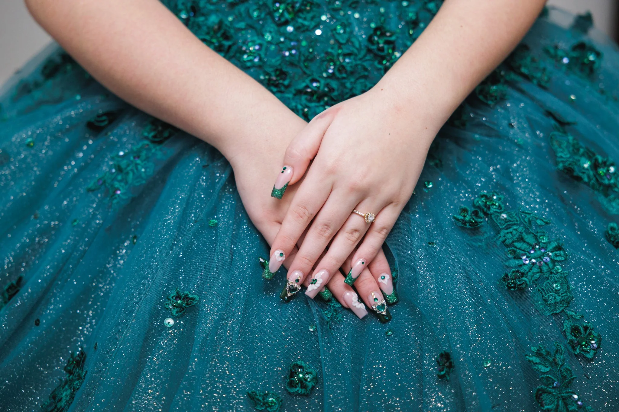 Close-up of a person's hands resting on a teal, sequined dress with floral and glitter embellishments. The person is wearing a diamond ring and has decorated fingernails with green glitter tips and small decorative elements.