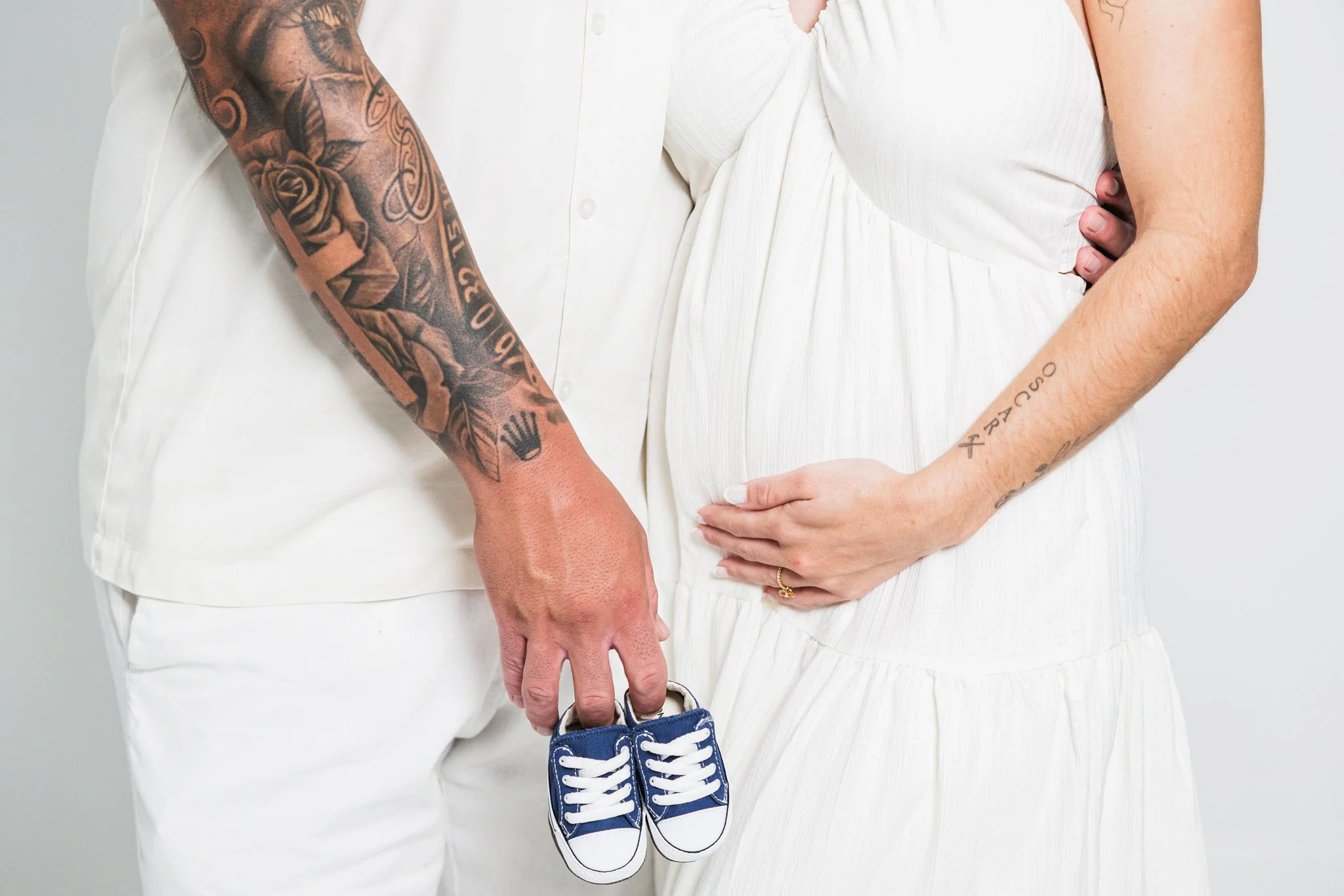 A pregnant woman in a white dress holding a pair of small blue sneakers. A man standing close by holding the woman's hand. The man has tattoos on his arm, the woman has tattoos on her arm.