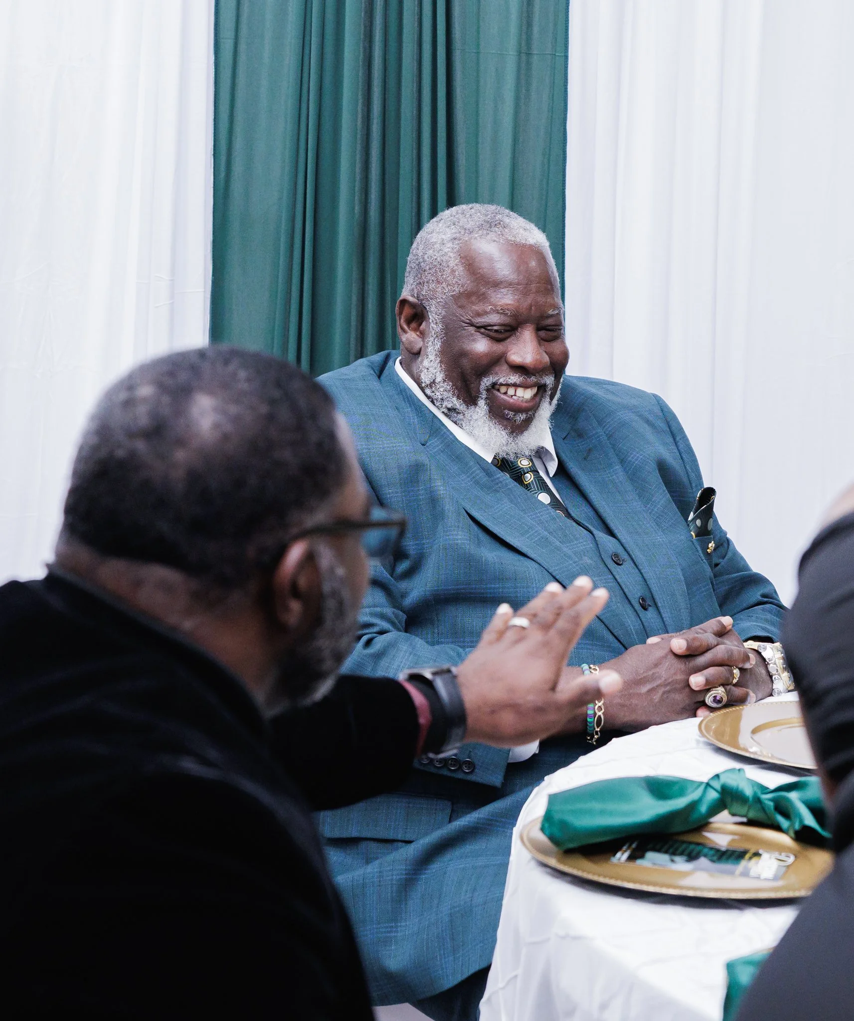A smiling man in a blue suit sitting at a dining table during a formal event, with two other men visible in the foreground.