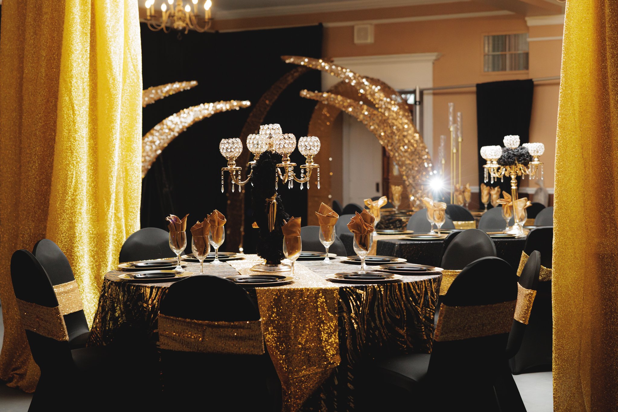 A decorated banquet table set with black, gold, and crystal accents, featuring ornate candelabras, champagne glasses with gold napkins, and gold sequined tablecloths, surrounded by black chairs with gold and sequined chair sashes, in a lavishly decor