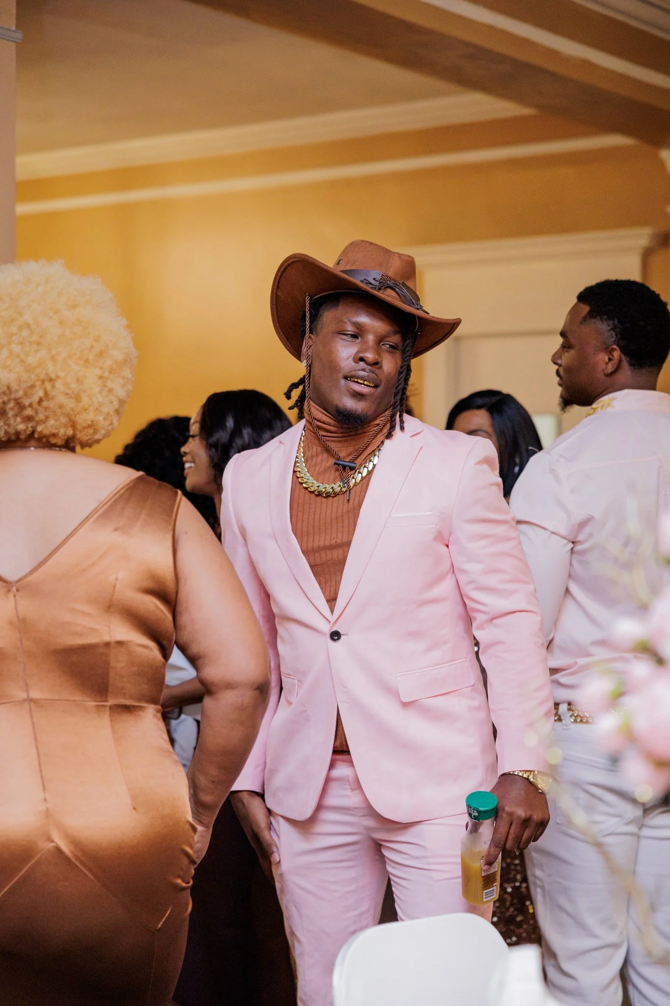 A man in a pink suit, brown turtleneck, gold chains, and a cowboy hat at a social gathering.