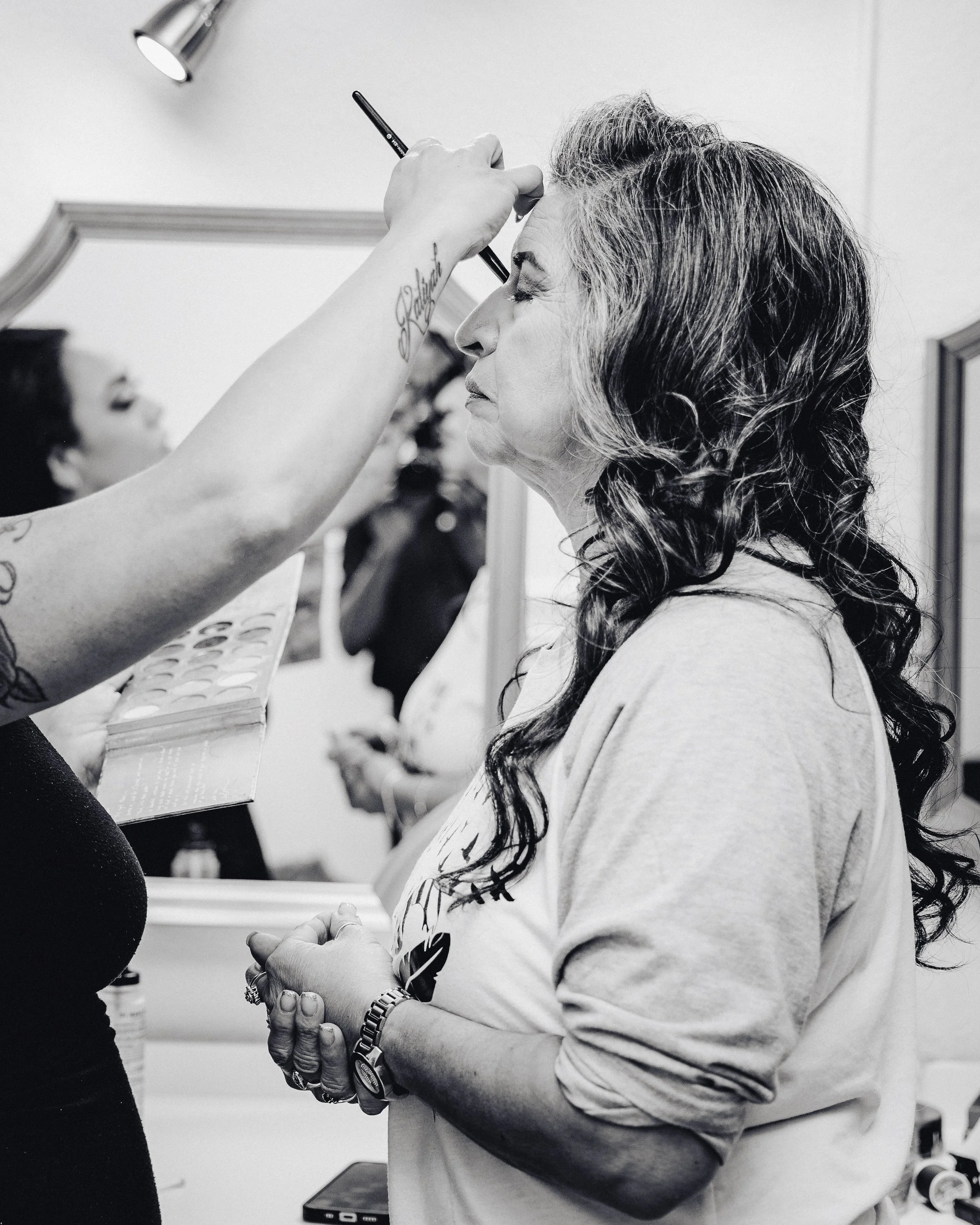 A woman getting her makeup done, with her eyes closed and hands clasped together, in a room with a mirror and another person in the background.