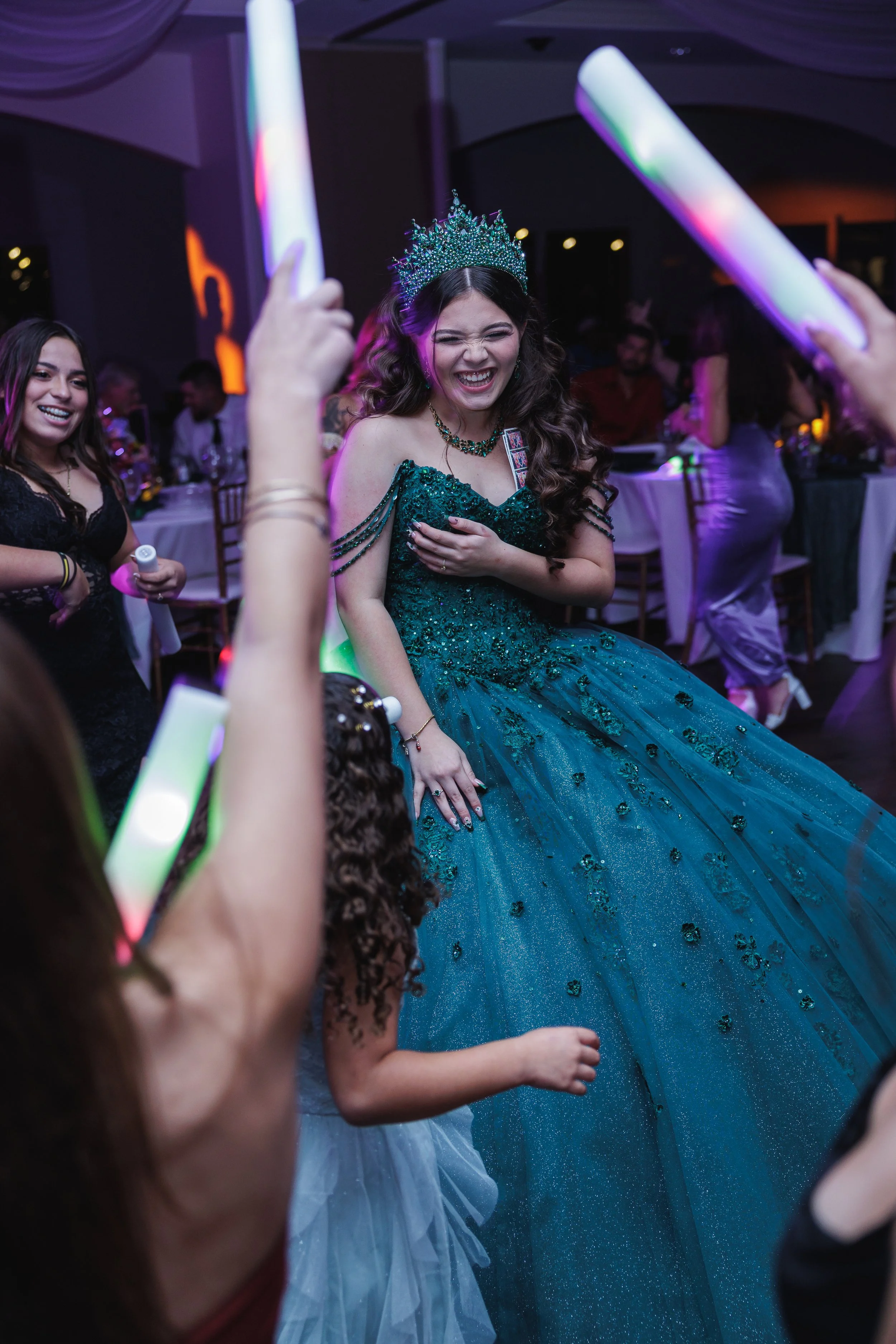 A woman in a teal, sparkly ball gown and crown laughs joyfully at a celebration, surrounded by friends holding glow sticks.
