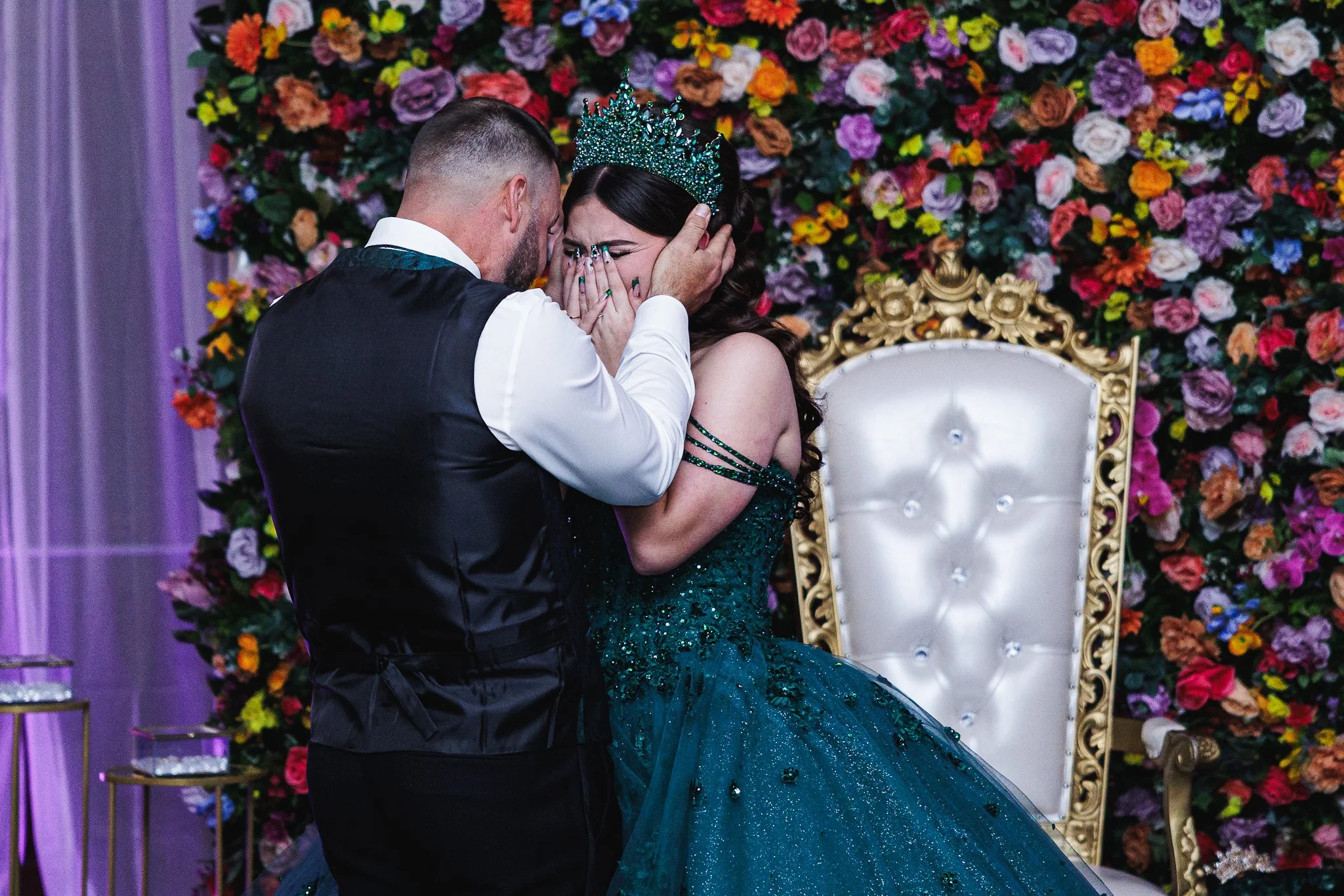 A bride wearing a green gown and a tiara is overwhelmed with emotion as a groom in a tuxedo comforts her at a wedding celebration, with a colorful floral backdrop and an ornate white and gold chair behind them.