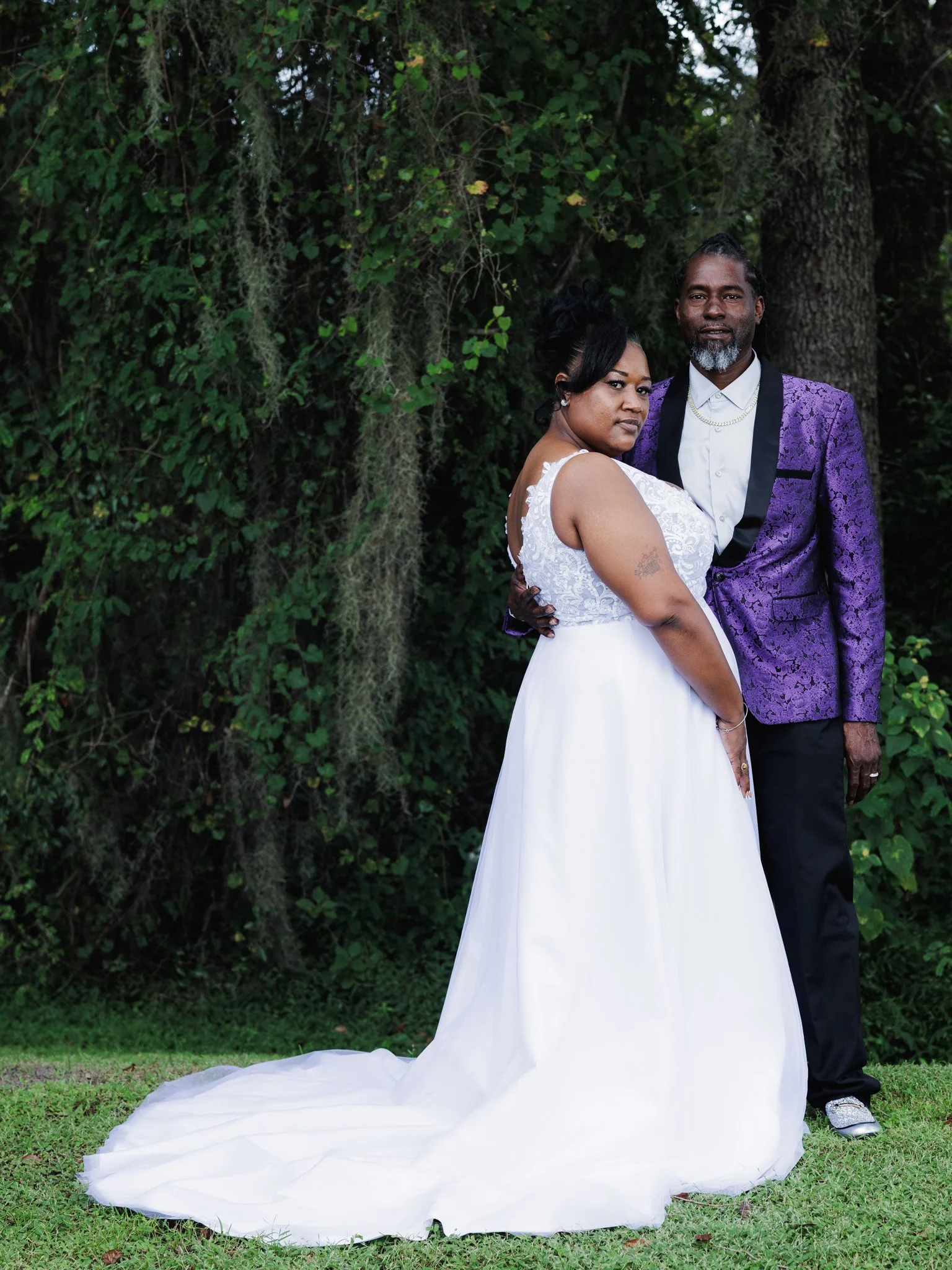 A bride in a white wedding dress and an older groom in a purple suit jacket stand together outdoors in front of green trees.