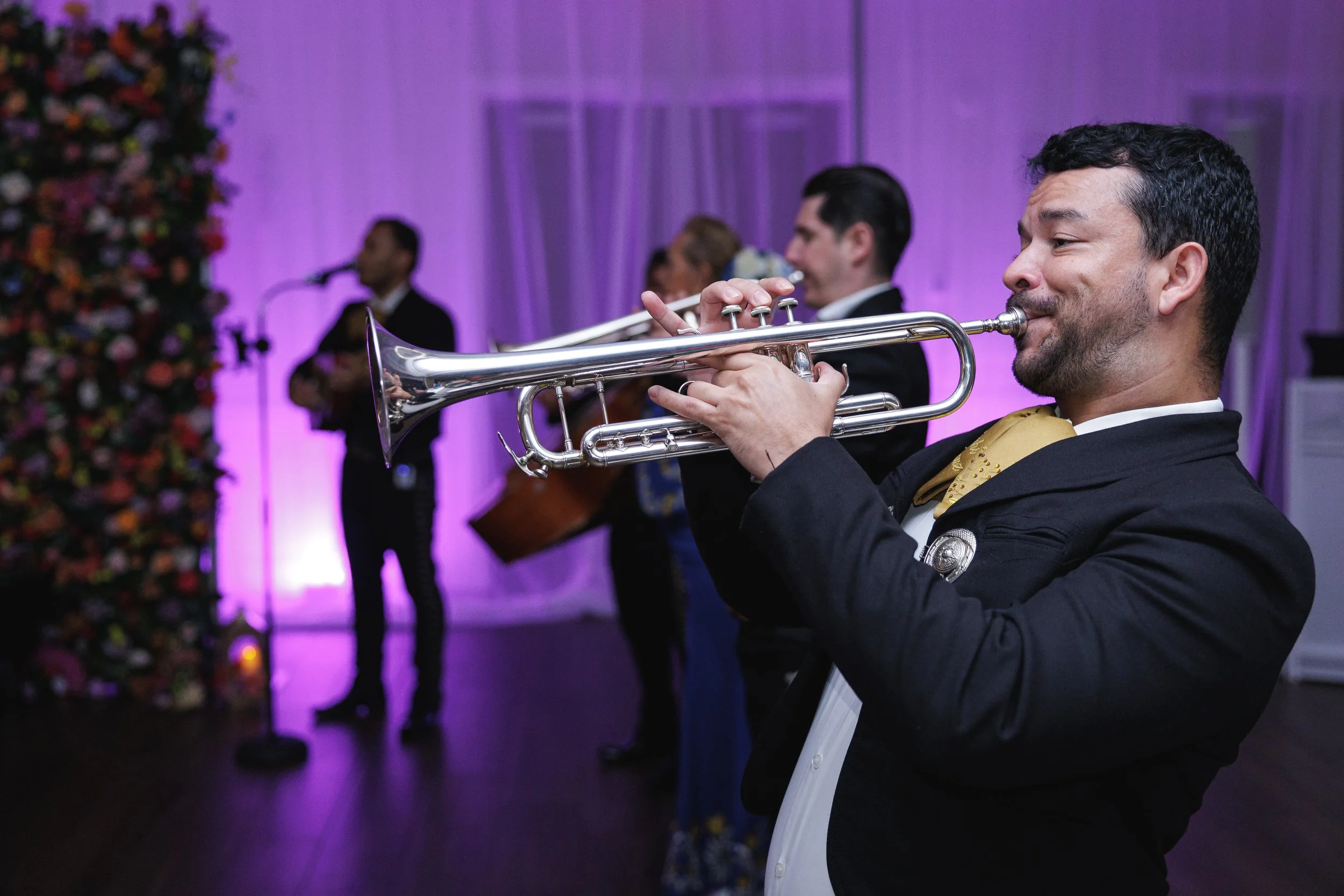 A man playing a trumpet at a festive event with other musicians performing in the background, decorated with purple lighting and a large flower arrangement.