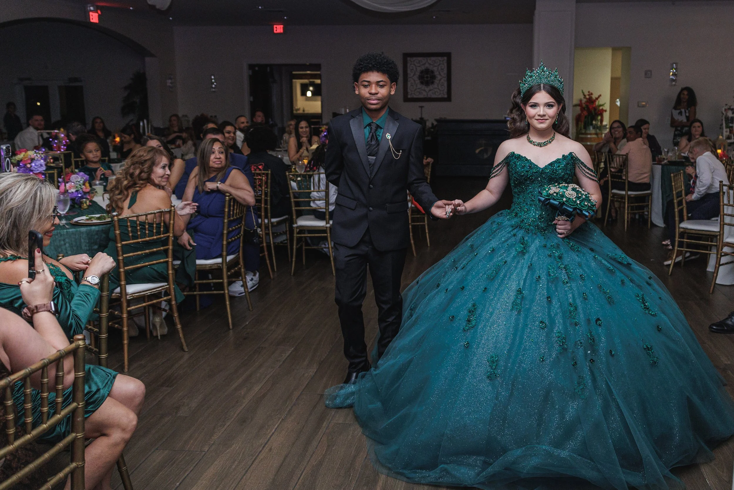 A young couple walking hand in hand during a formal event, with the girl wearing an elaborate green gown and crown, and the boy in a dark suit, surrounded by seated guests at banquet tables.