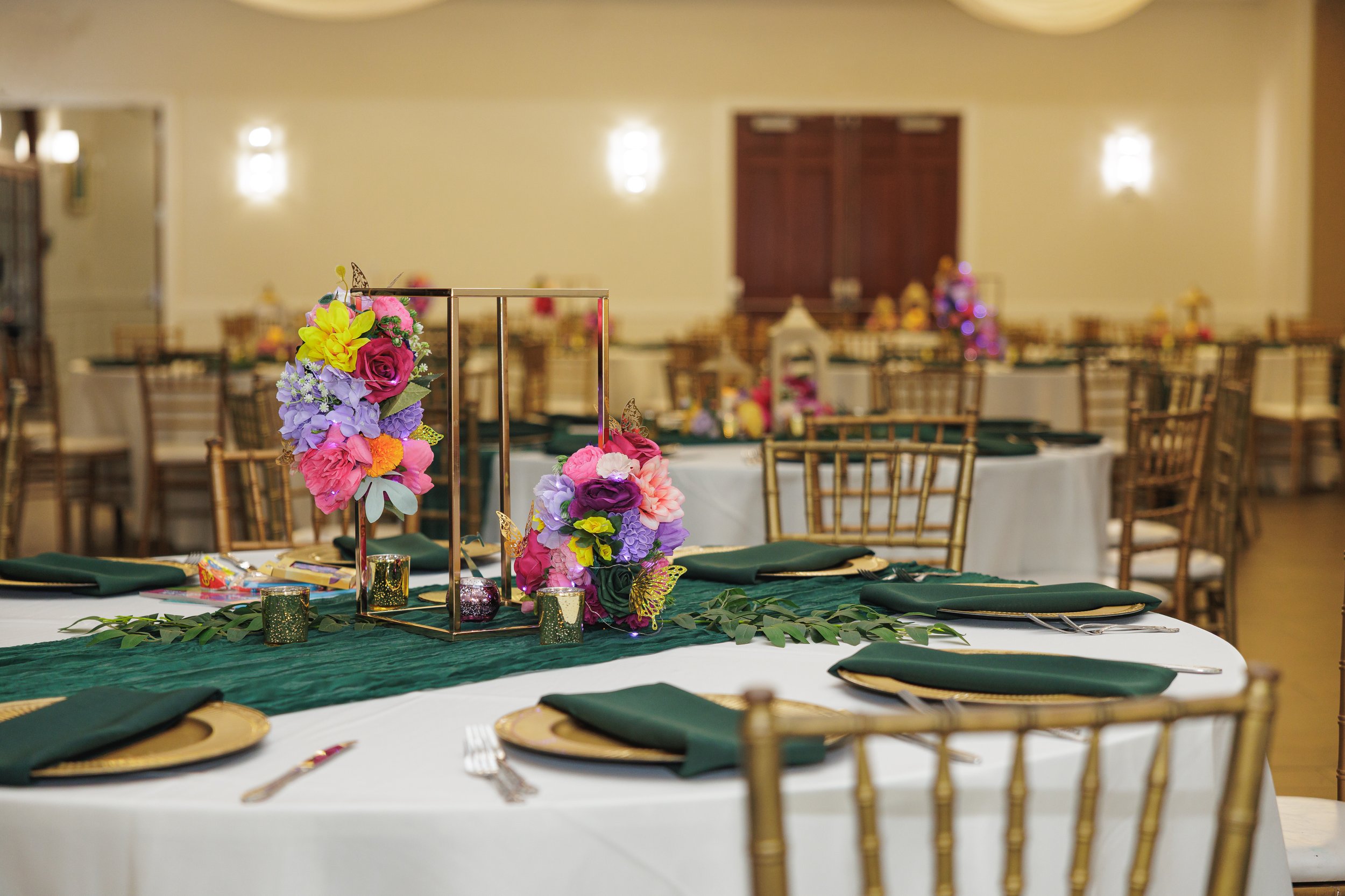 A banquet hall decorated for a celebration with round tables, green napkins, gold chargers, and floral centerpieces featuring pink, purple, and yellow flowers.