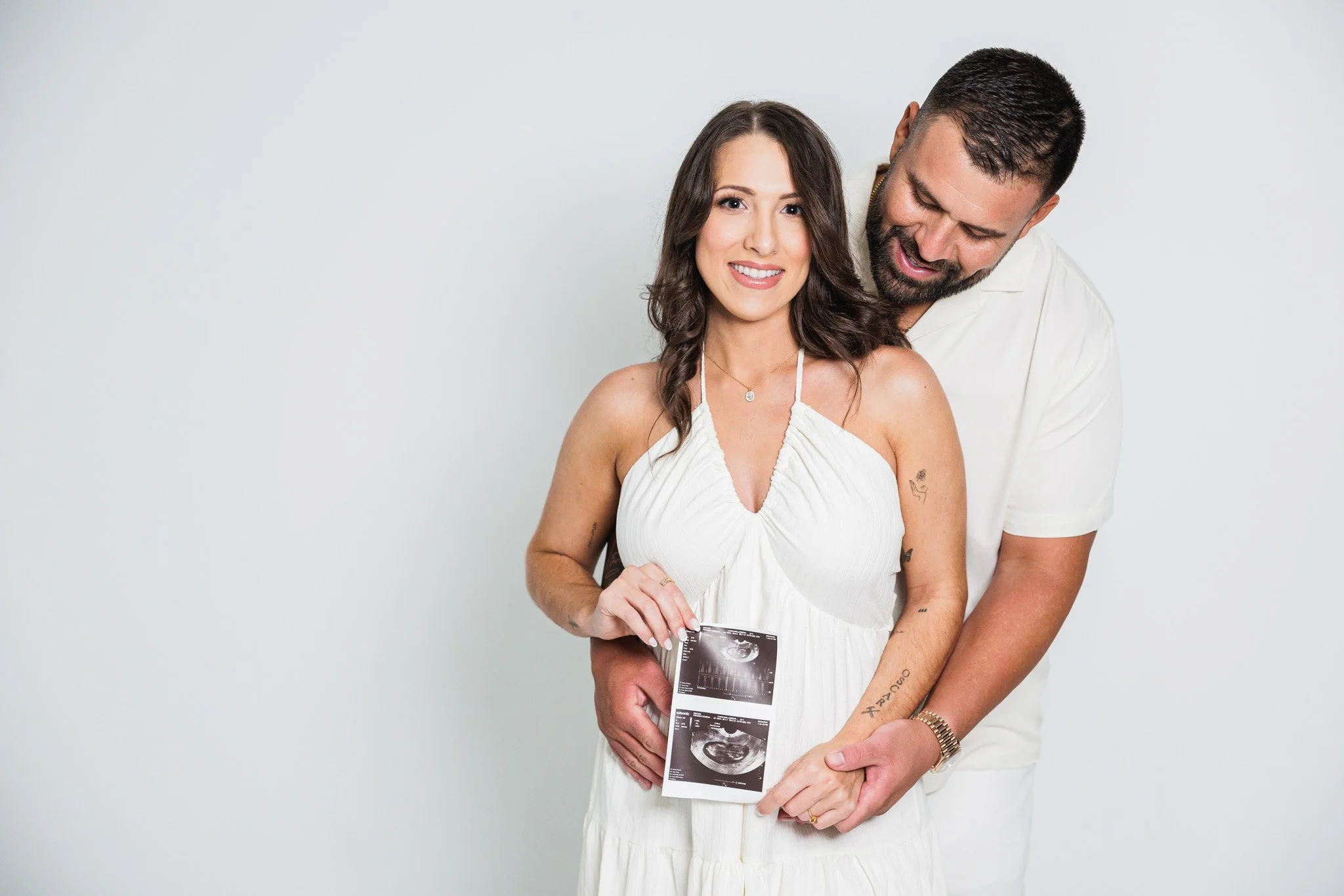 A smiling woman holding ultrasound pictures, with a man standing behind her, wrapping his arms around her waist, in a light-colored room.