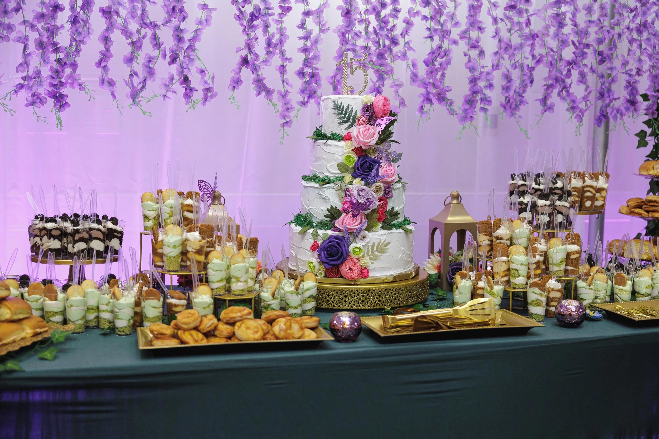 A decorated celebration table with a multi-tiered white cake adorned with purple, pink, and white roses, and purple butterflies, surrounded by an assortment of desserts including petit fours, cookies, and parfaits in small cups, all set against a pur